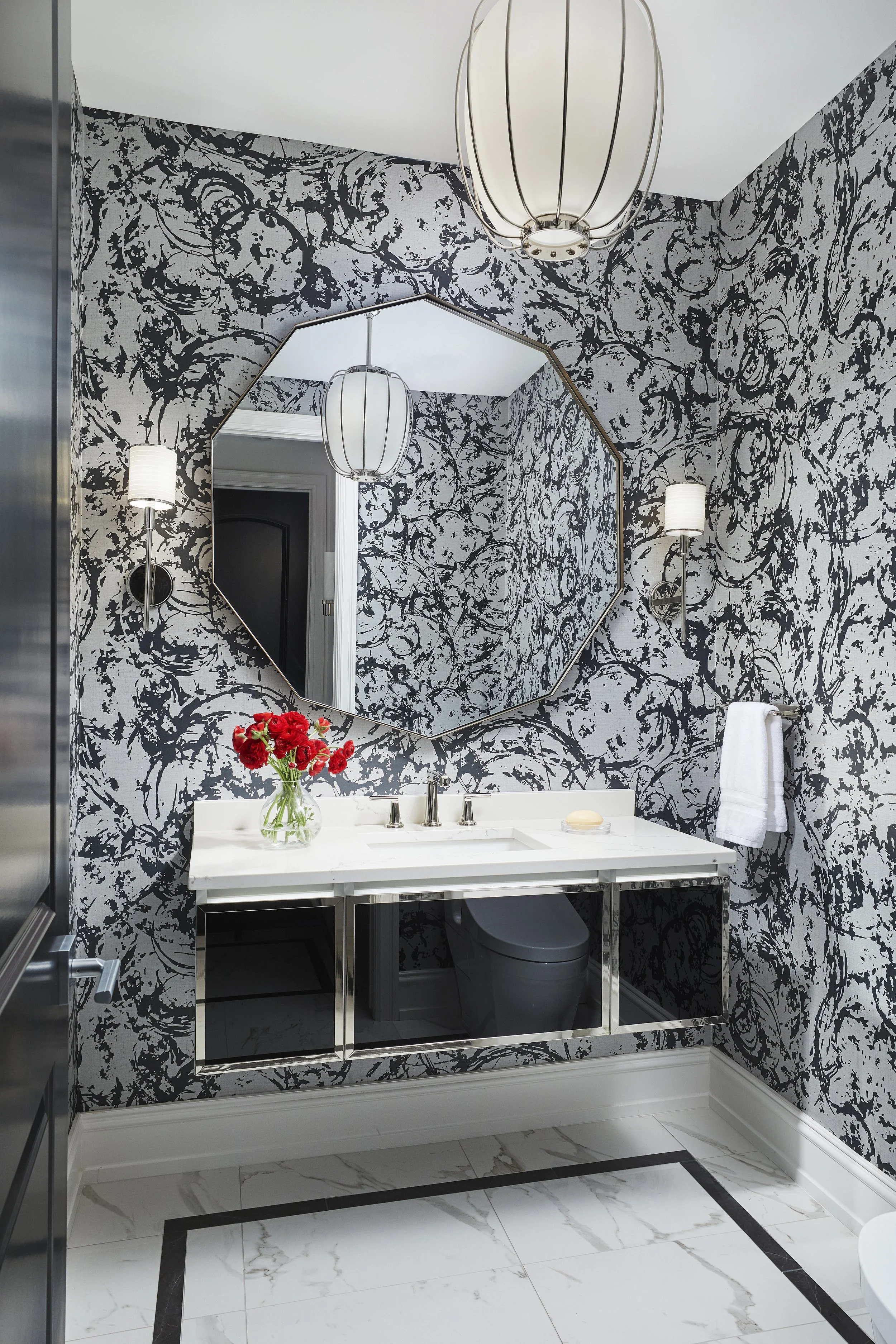 A bathroom with black and white marble patterned wallpaper, a white sink with a flower vase on top, a large octagonal mirror, wall-mounted lights, a towel next to the sink, and black and white marble floor tiles.