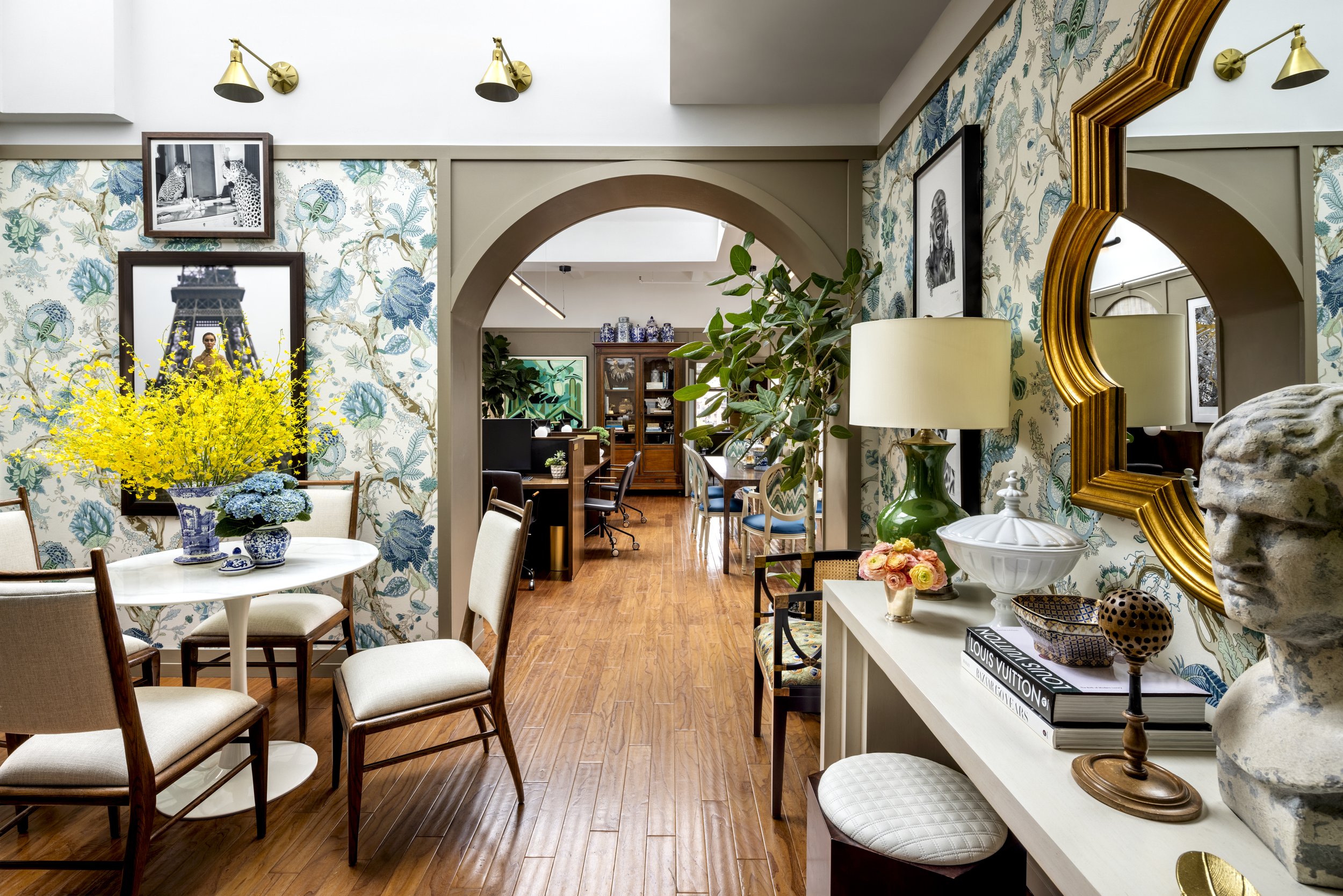 Interior of a stylish room with floral wallpaper, wooden flooring, a round table with white chairs, various decorative items, a large yellow flower arrangement, and a mirror with a gold frame.
