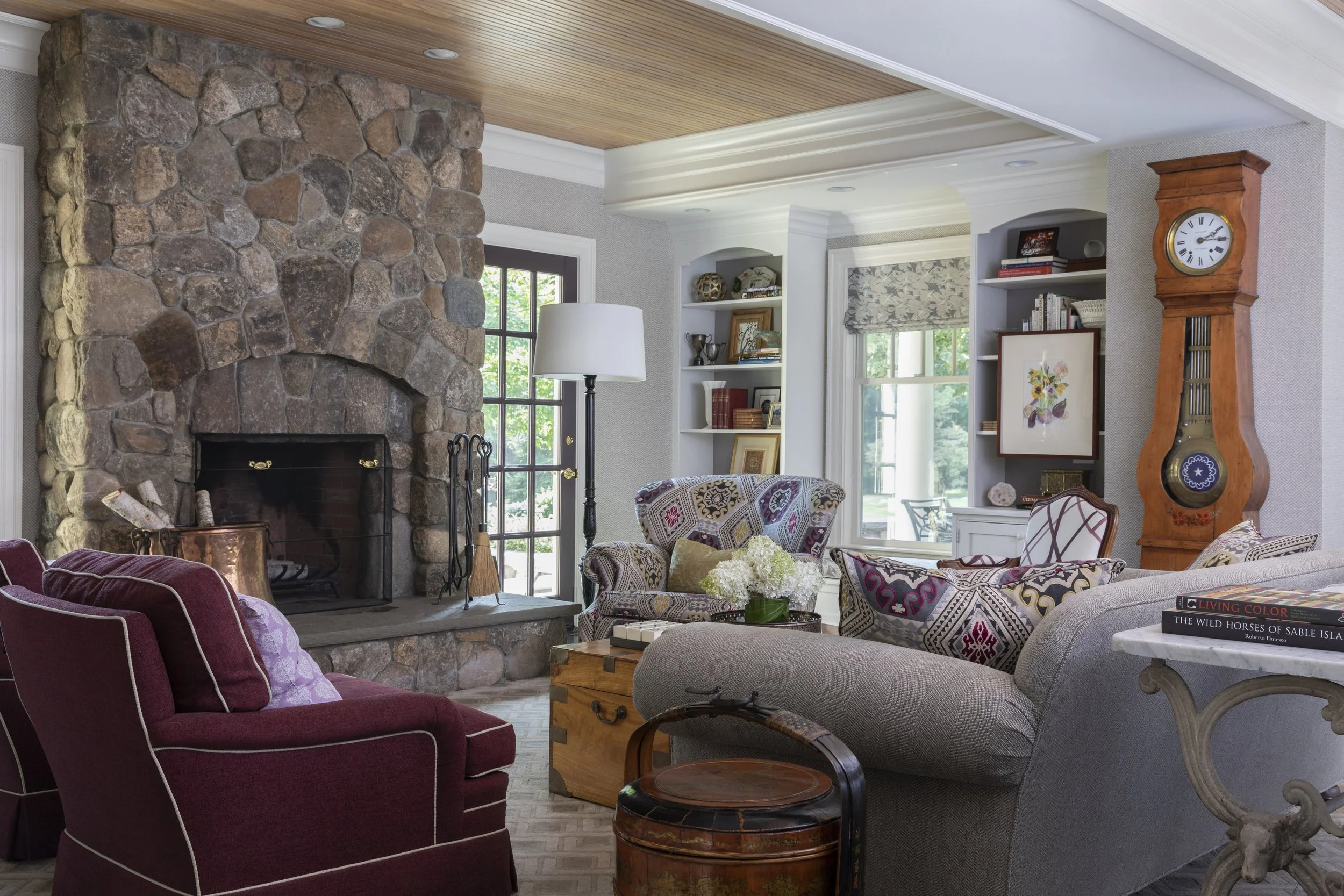 Living room with a large stone fireplace, patterned upholstered chairs, a white couch with pillows, a wooden coffee table, a bookshelf, a tall wooden grandfather clock, and large windows with views of greenery.