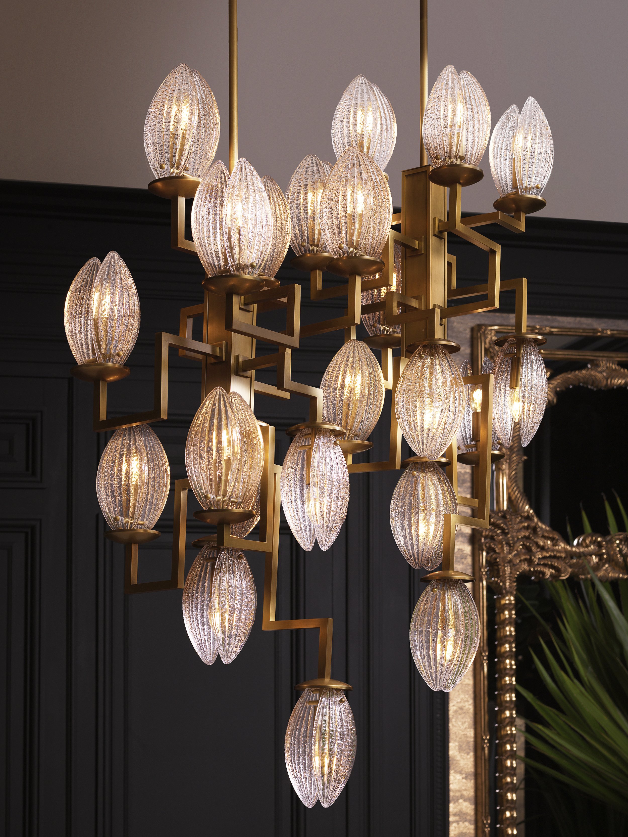 Elegant chandelier with multiple glass or crystal elongated oval-shaped shades emitting warm light, mounted in a room with dark walls and decorative elements.