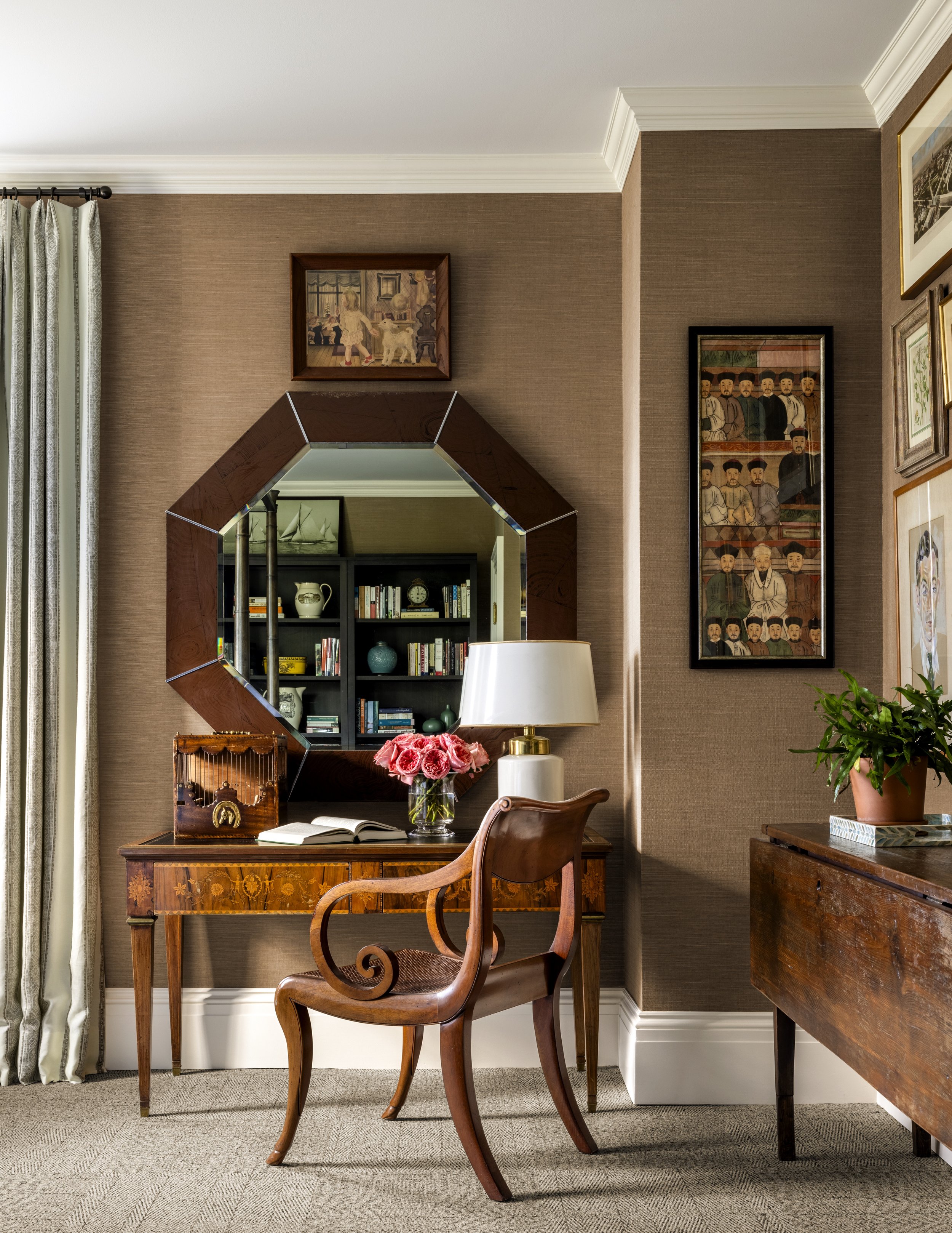 A cozy home office with a wooden desk, a carved wooden chair, decorative framed artwork on brown textured walls, a hexagonal mirror, a table lamp, a vase of pink roses, a bookshelf, and a potted plant.
