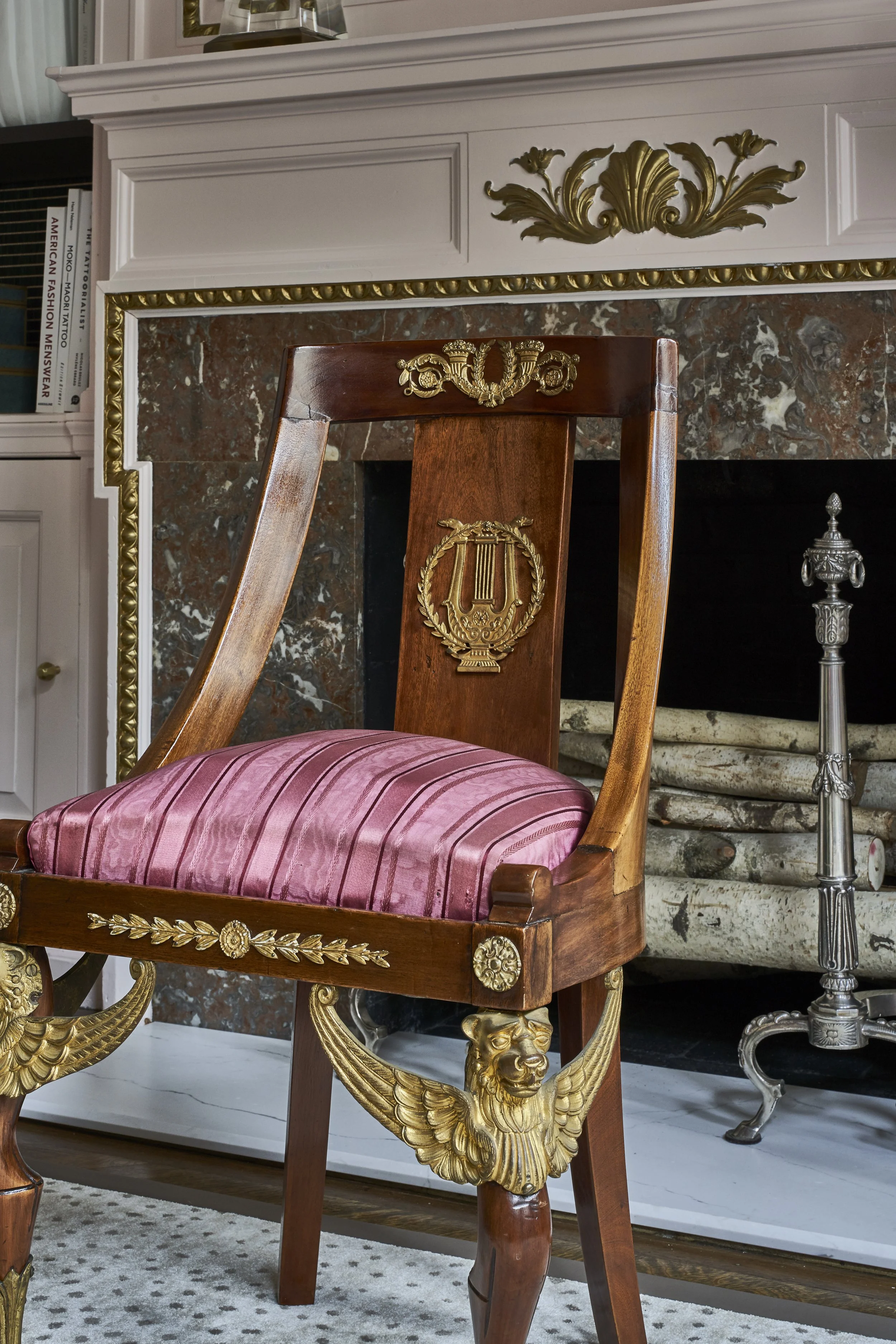 An ornate wooden chair with gold accents and a pink striped cushion, positioned in front of a fireplace with a marble hearth and decorative molding.
