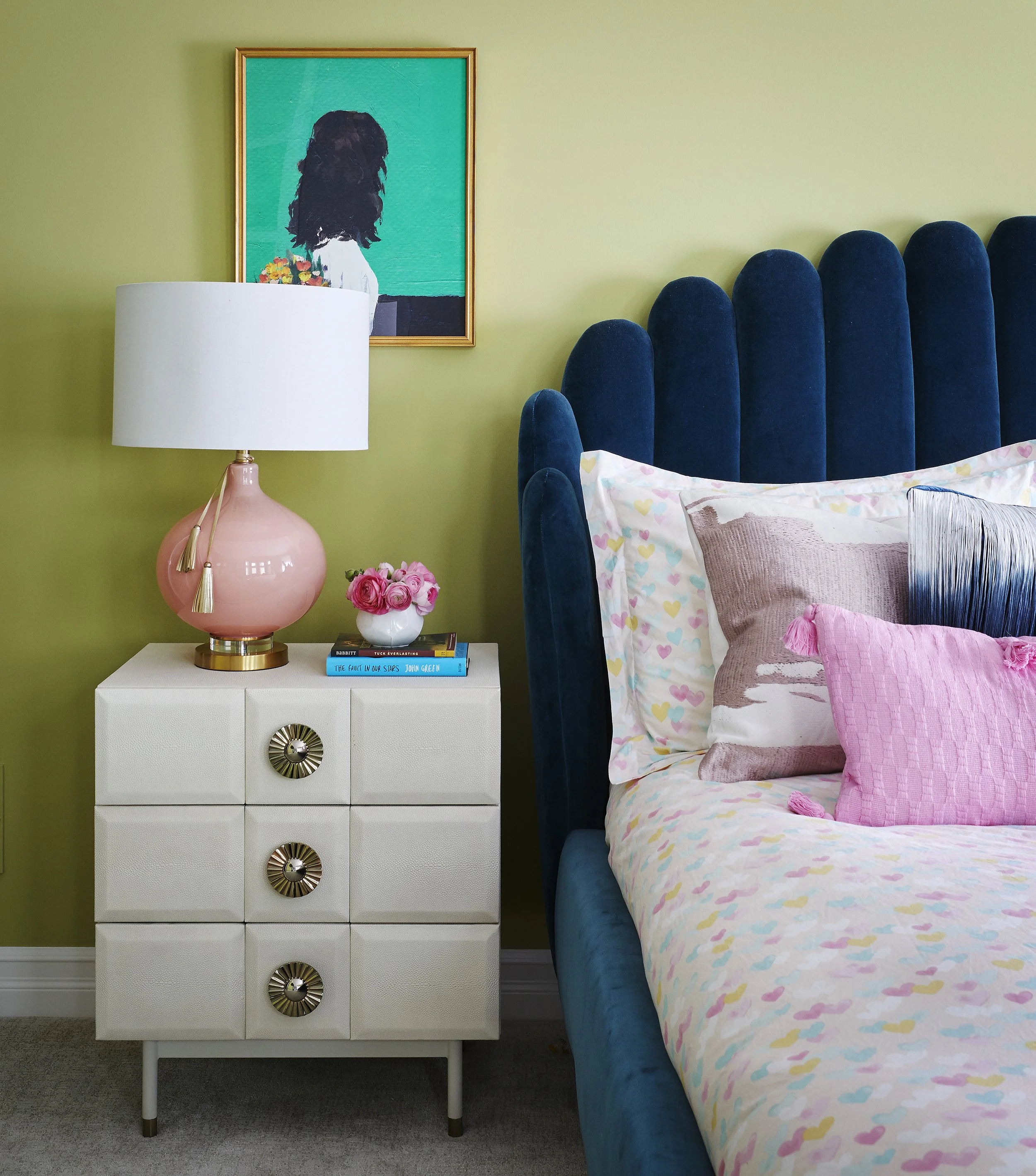 A bedroom with a green wall, blue velvet headboard, white side table with a pink lamp, a stack of books, pink flowers, and a bed with pastel-colored heart-patterned sheets and pink and beige pillows. There is a framed portrait of a girl with dark hai