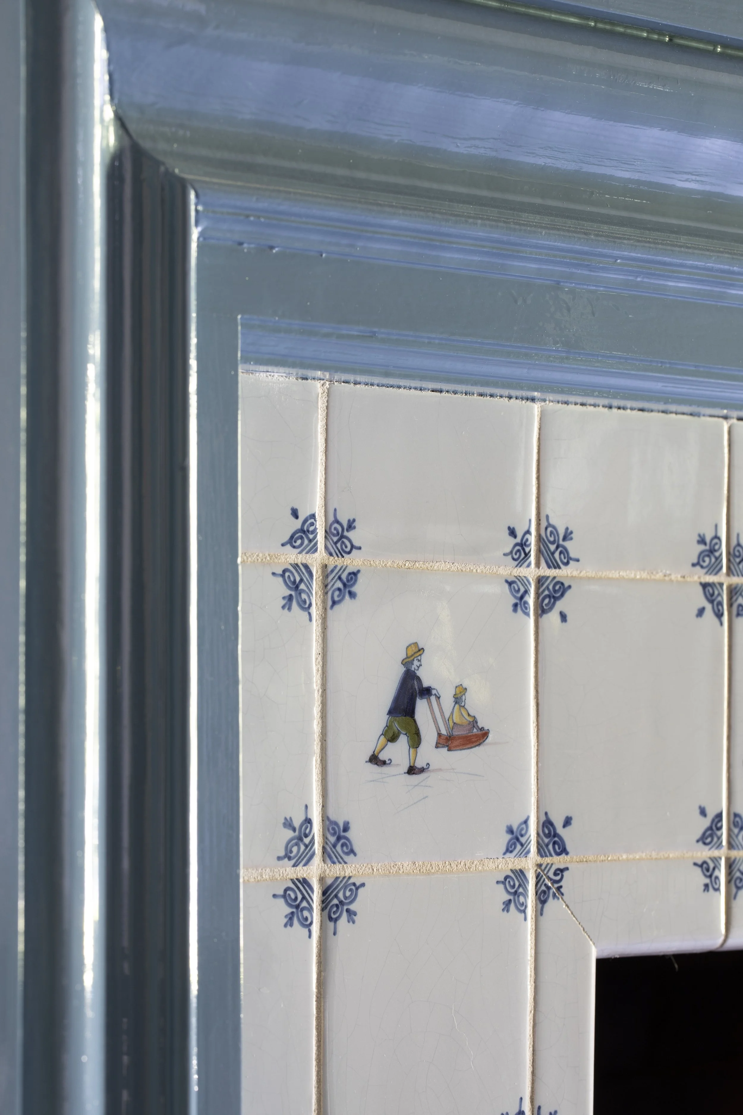 Close-up of tiled wall with decorative blue patterns, partially obscured by window frame on the left. The wall features illustration of two individuals in yellow hats, one pushing a stroller, seen through the window. The scene appears to be inside a 
