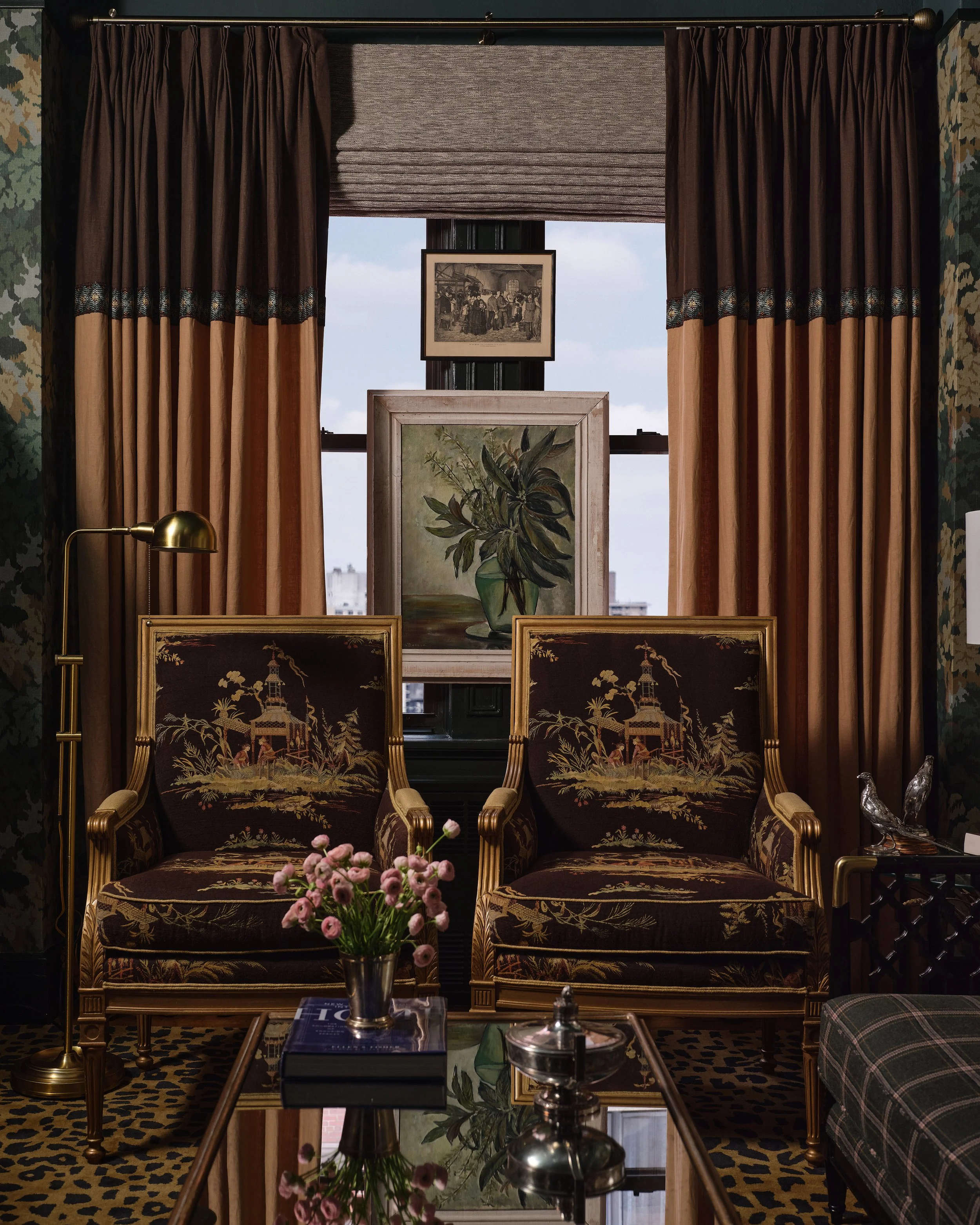 A room with two ornate chairs featuring embroidered scenes of pagodas and trees, a side table with a pink flower arrangement, a glass-topped coffee table with books and silver decorative objects, and a window with brown and beige curtains and a rolle