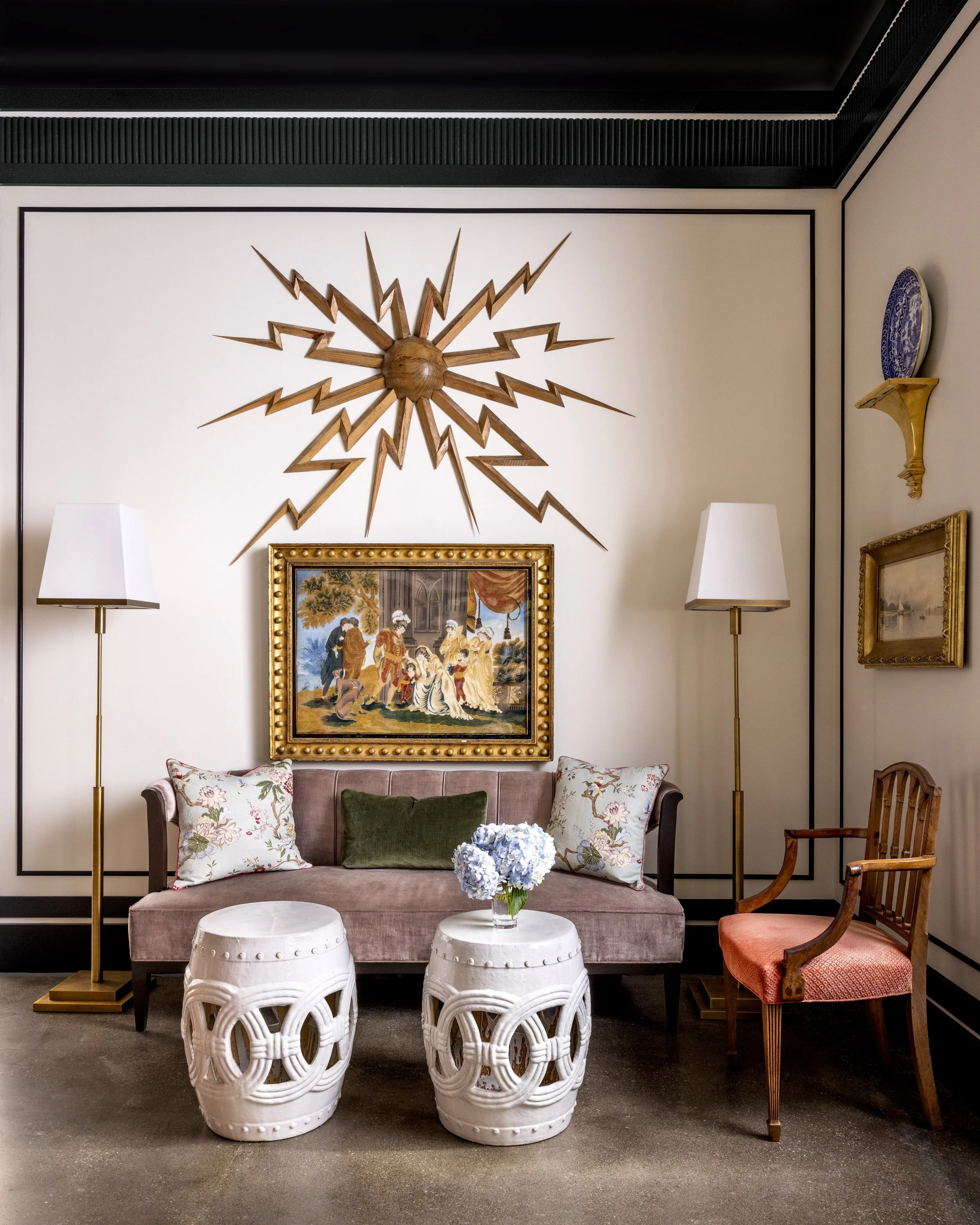 A cozy living room corner with beige walls and black trim. A mauve velvet sofa with floral and green pillows is in front of a framed vintage painting. Two white sculptural side tables with organic shapes hold a glass vase with blue hydrangeas. Two ta