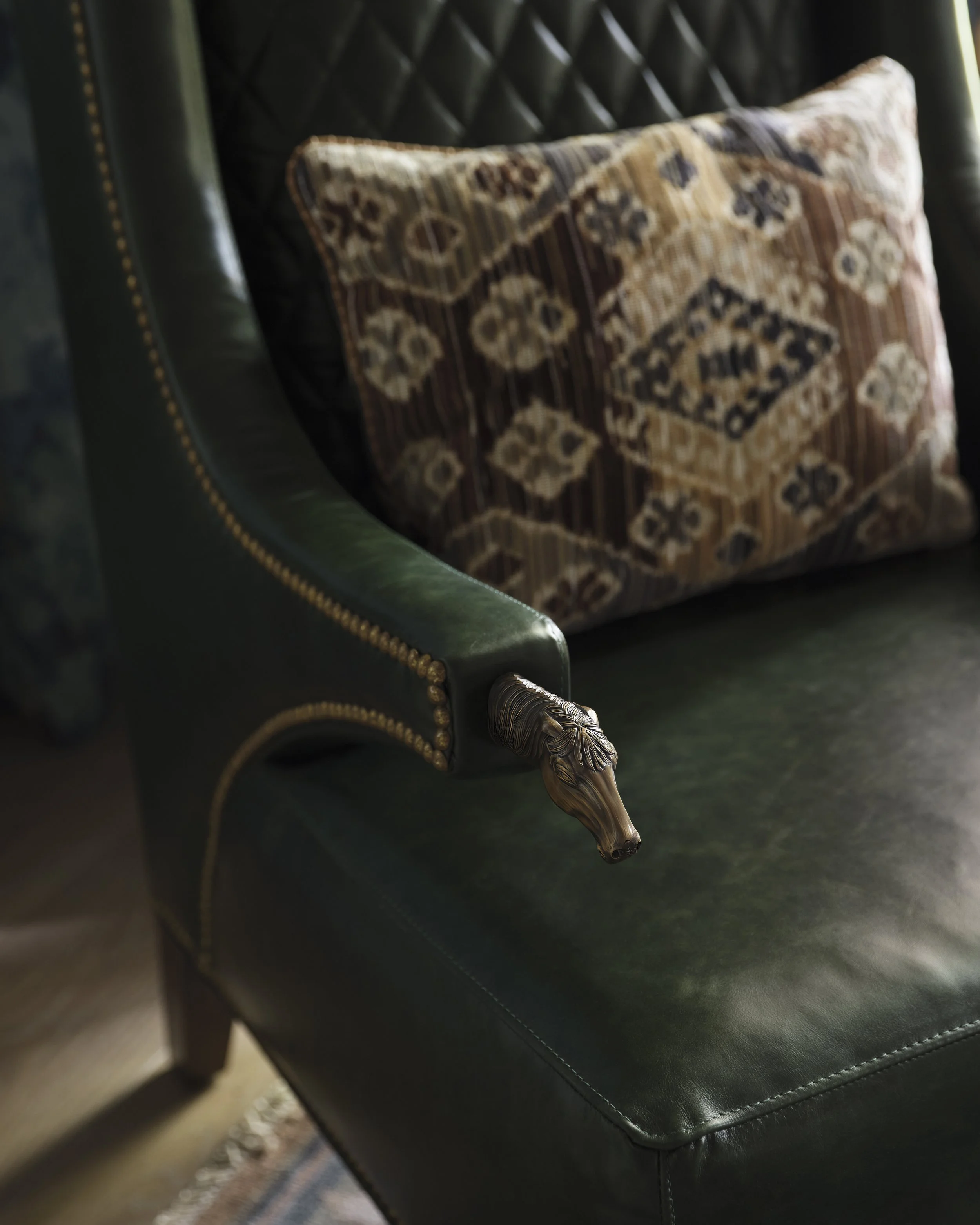 An ornate armchair with green leather upholstery, brass nailhead trim, and decorative wood carving on the armrest, situated against a dark backdrop.