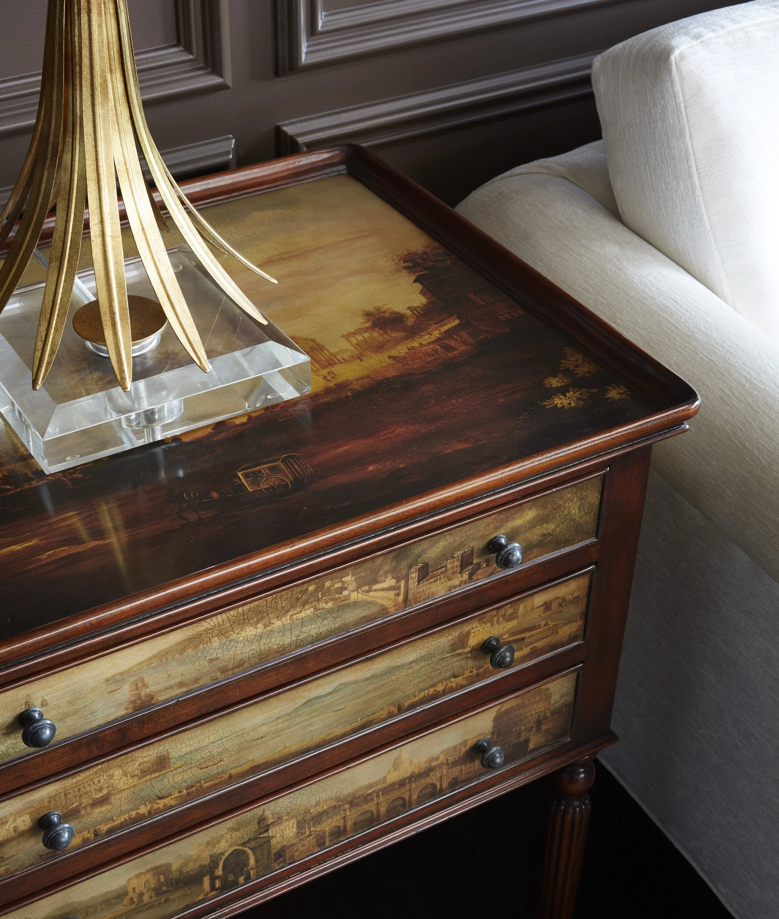 Close-up of a wooden dresser with scenic landscape paintings on the top surfaces of the drawers, a brass decorative lamp with elongated leaves, and a light-colored upholstered couch with a rolled armrest in a well-decorated room.