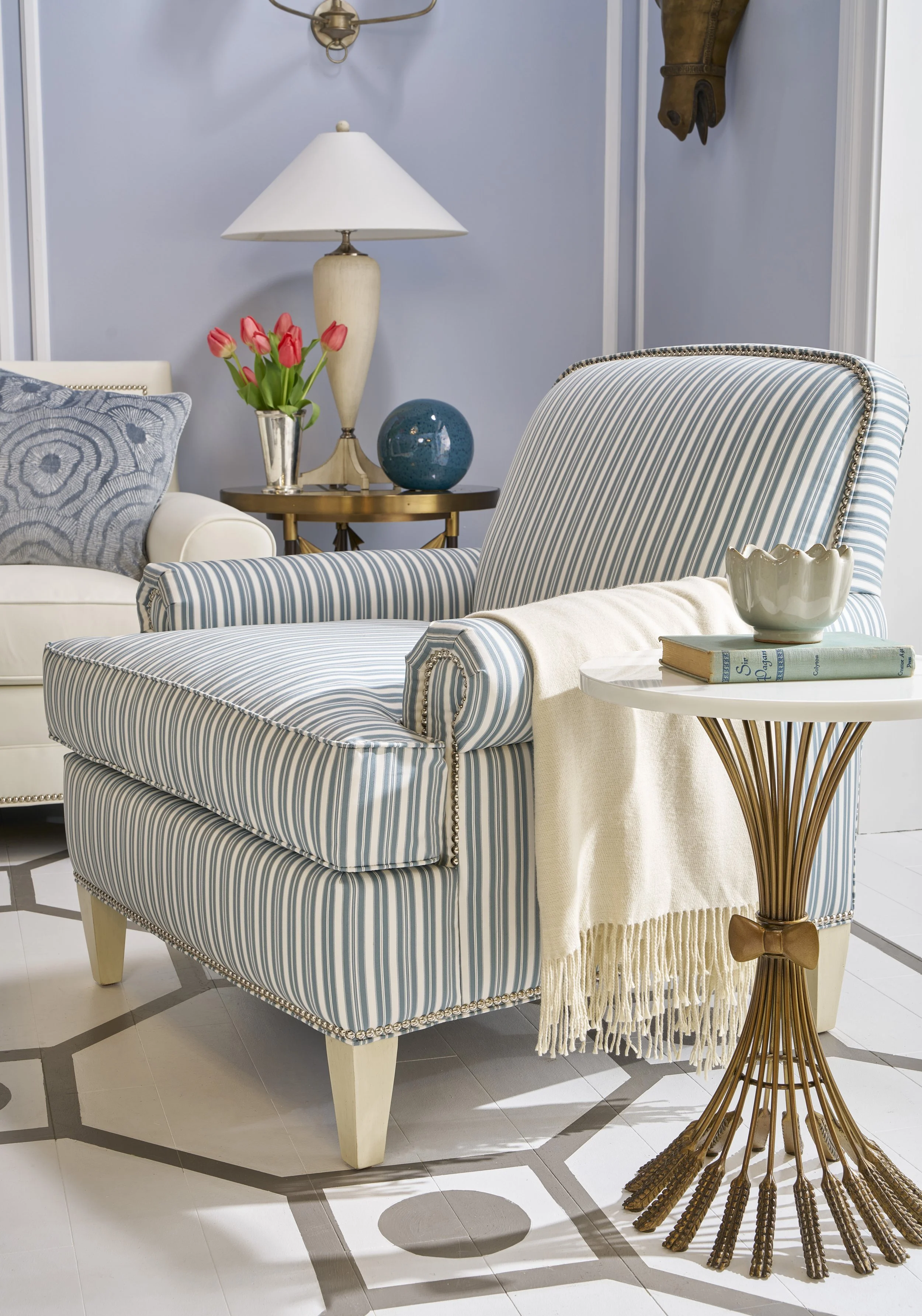 A living room scene with a blue and white striped armchair and a white sofa with a blue patterned pillow. There is a round side table with a vase of pink tulips, a blue decorative orb, and a lamp with a beige base and white shade. A fringed throw bla