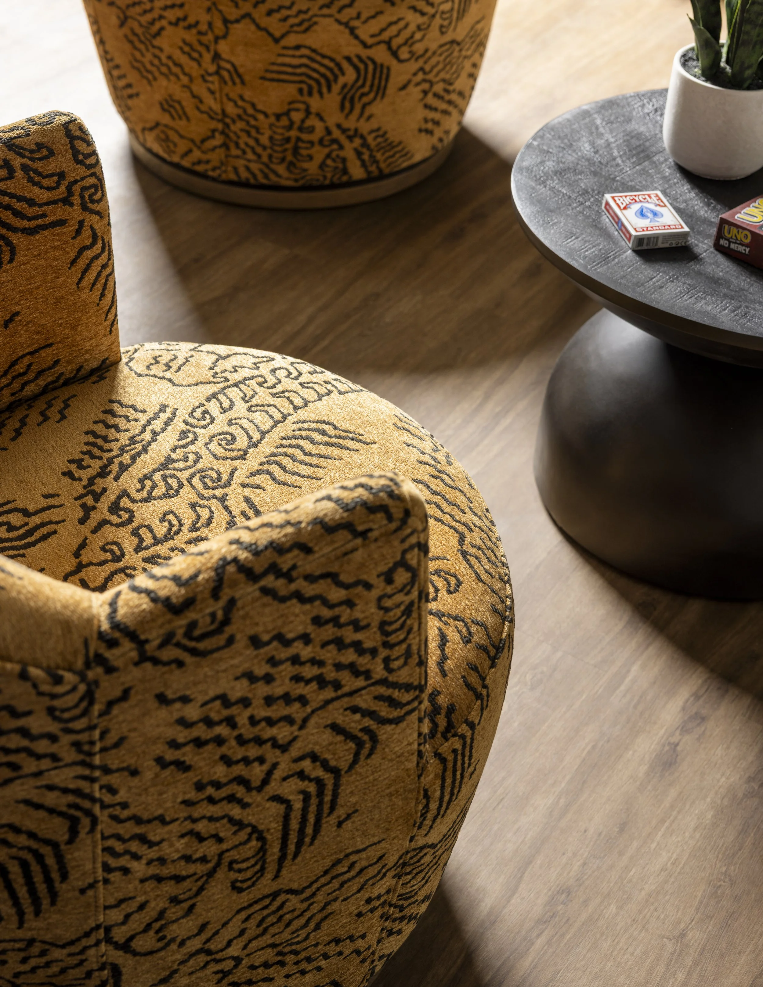 Close-up view of a rounded, cushioned armchair with yellow velvet upholstery featuring black tiger-like stripes. A small round dark wooden table is next to the armchair, with a white pot containing a green succulent and two small boxes of games, Uno 