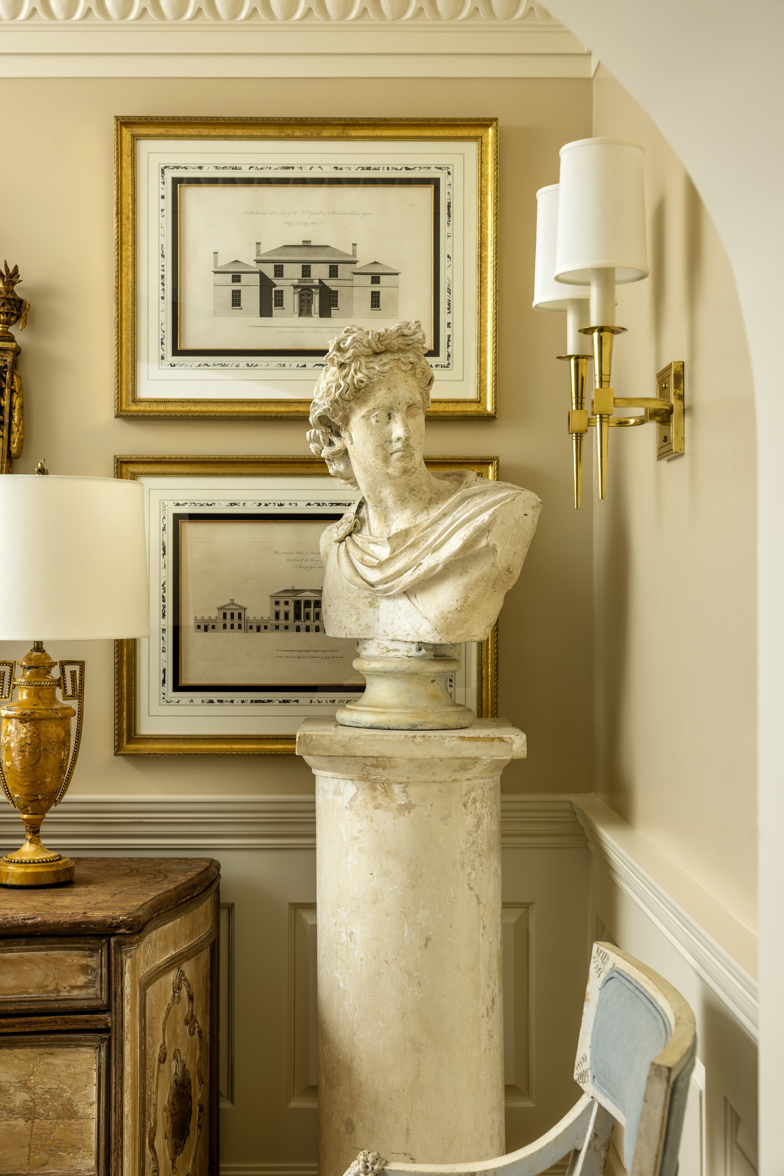 A marble bust sculpture of a woman's head and shoulders on a pedestal, behind a light-colored wall with framed architectural drawings and a wall-mounted lamp with gold accents, in an elegant interior setting.