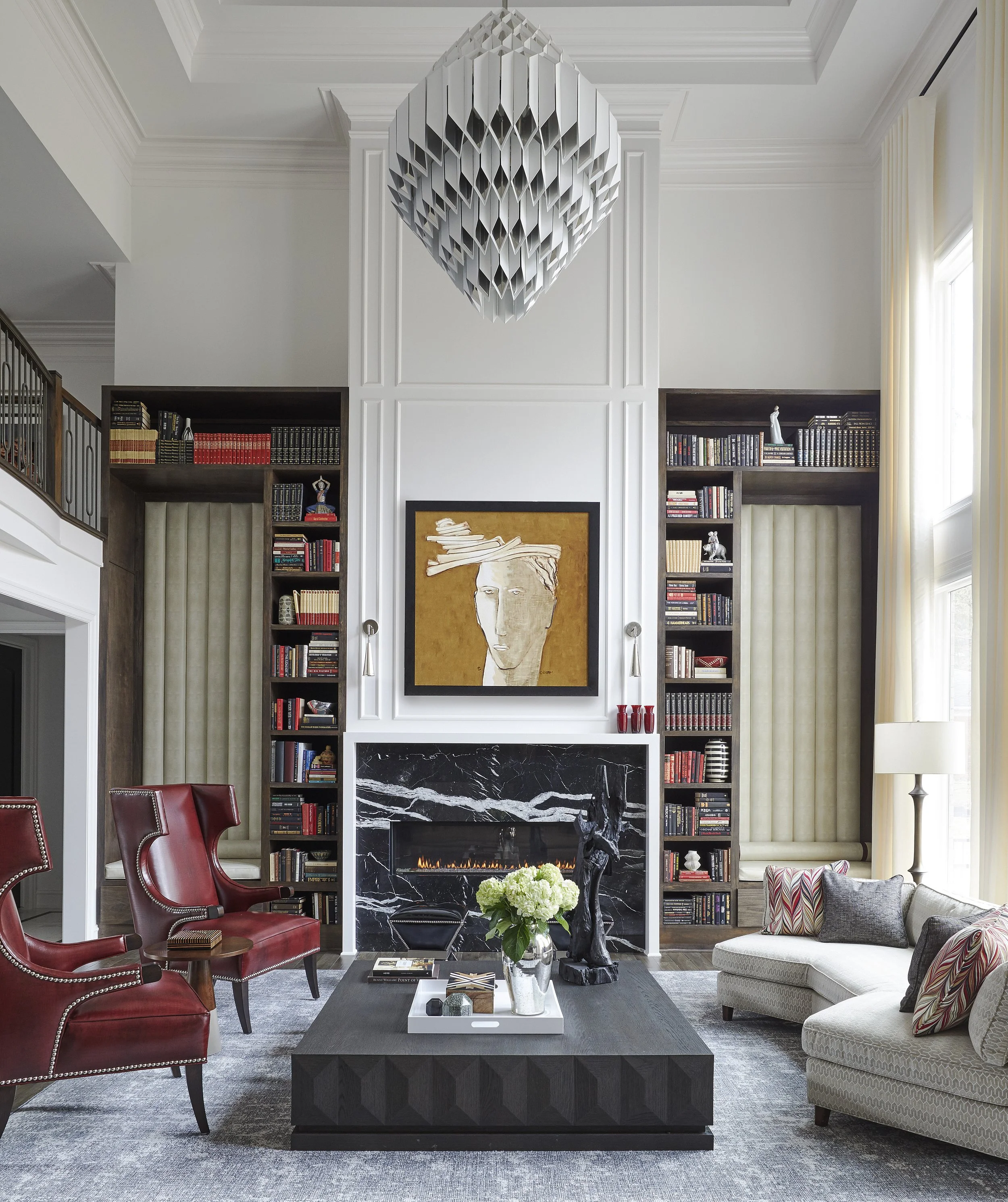 Living room with fireplace, black marble surround, bookshelf on either side, artwork above mantel, red and patterned armchairs, beige sofa with pillows, large window with curtains, chandelier, flowers on coffee table.