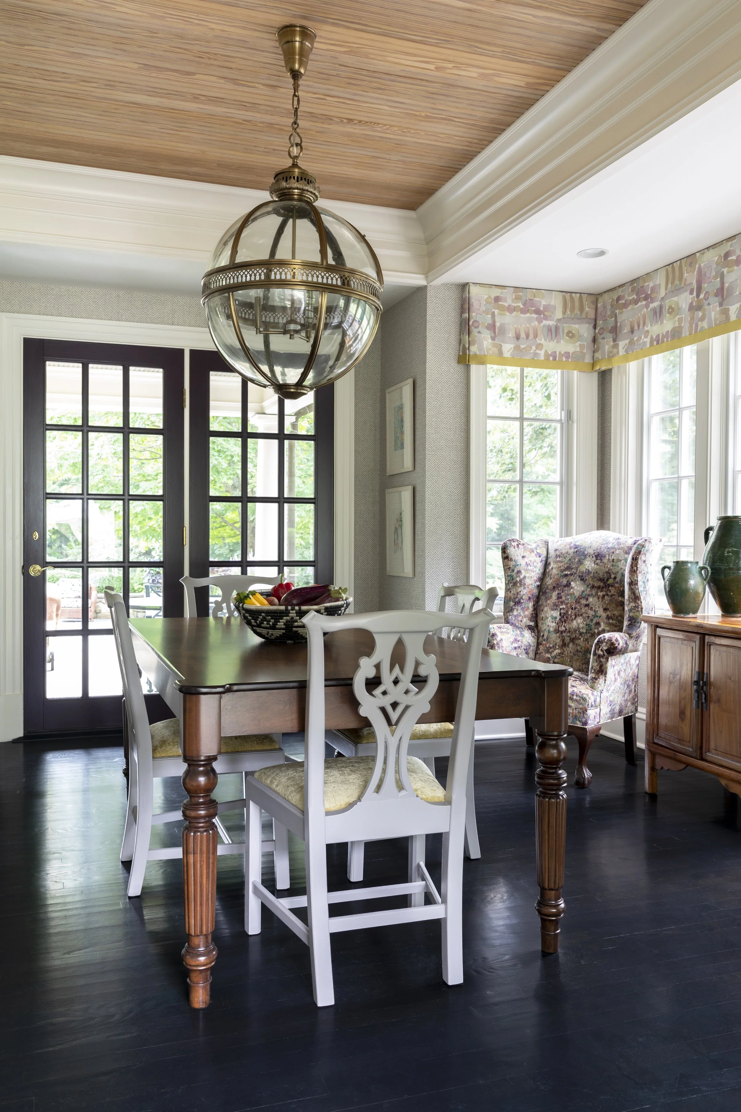 A dining room with a wooden table, white chairs, a fruit bowl, large windows, a vintage chandelier, patterned curtains, and an armchair with a floral cover.