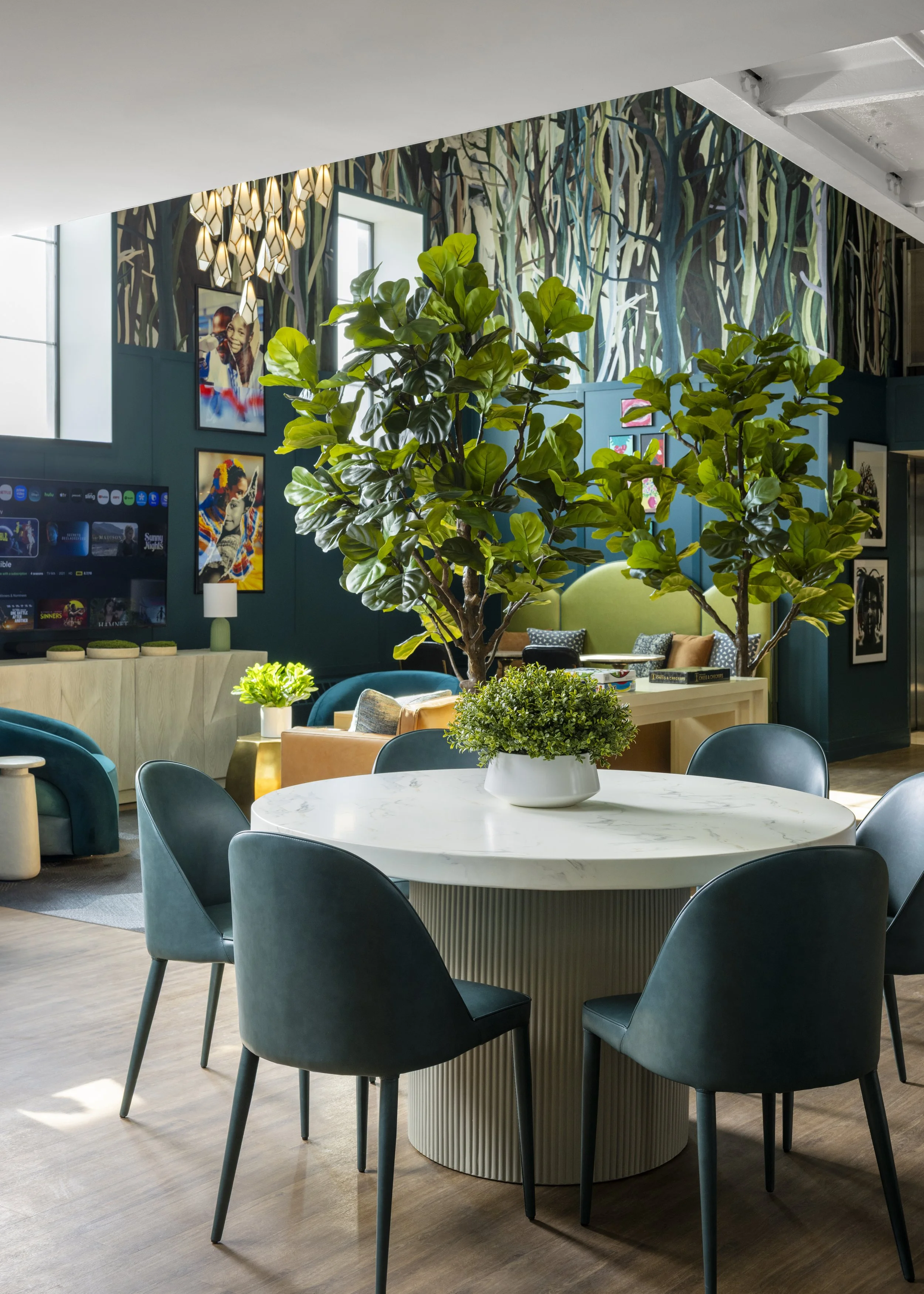 Modern indoor lounge area with a round table, dark blue chairs, potted plants, wall art, and hanging light fixtures.
