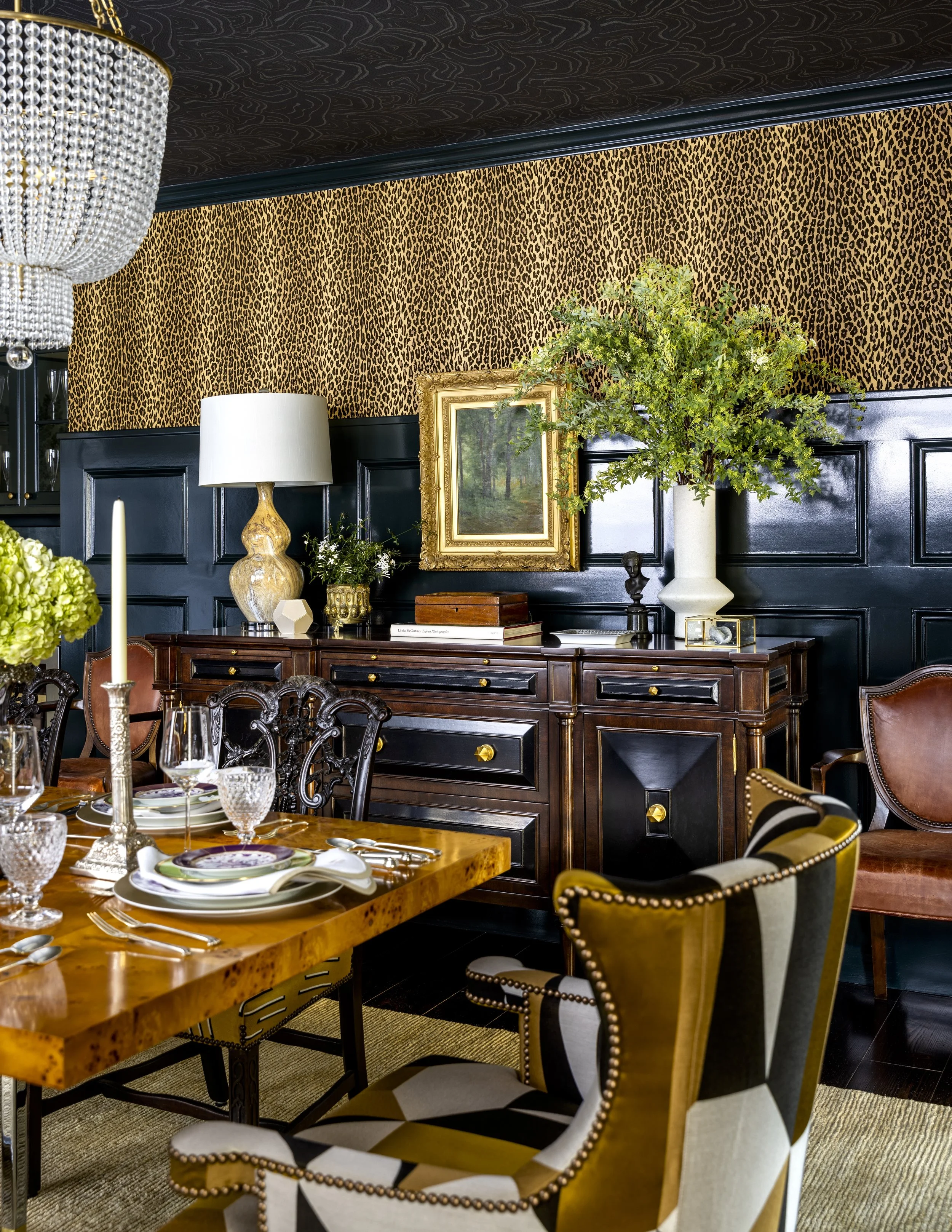 Interior of a dining room with a leopard print patterned wallpaper, a dark wood sideboard, a tall white vase with green foliage, a gold-framed landscape painting, and a crystal chandelier.