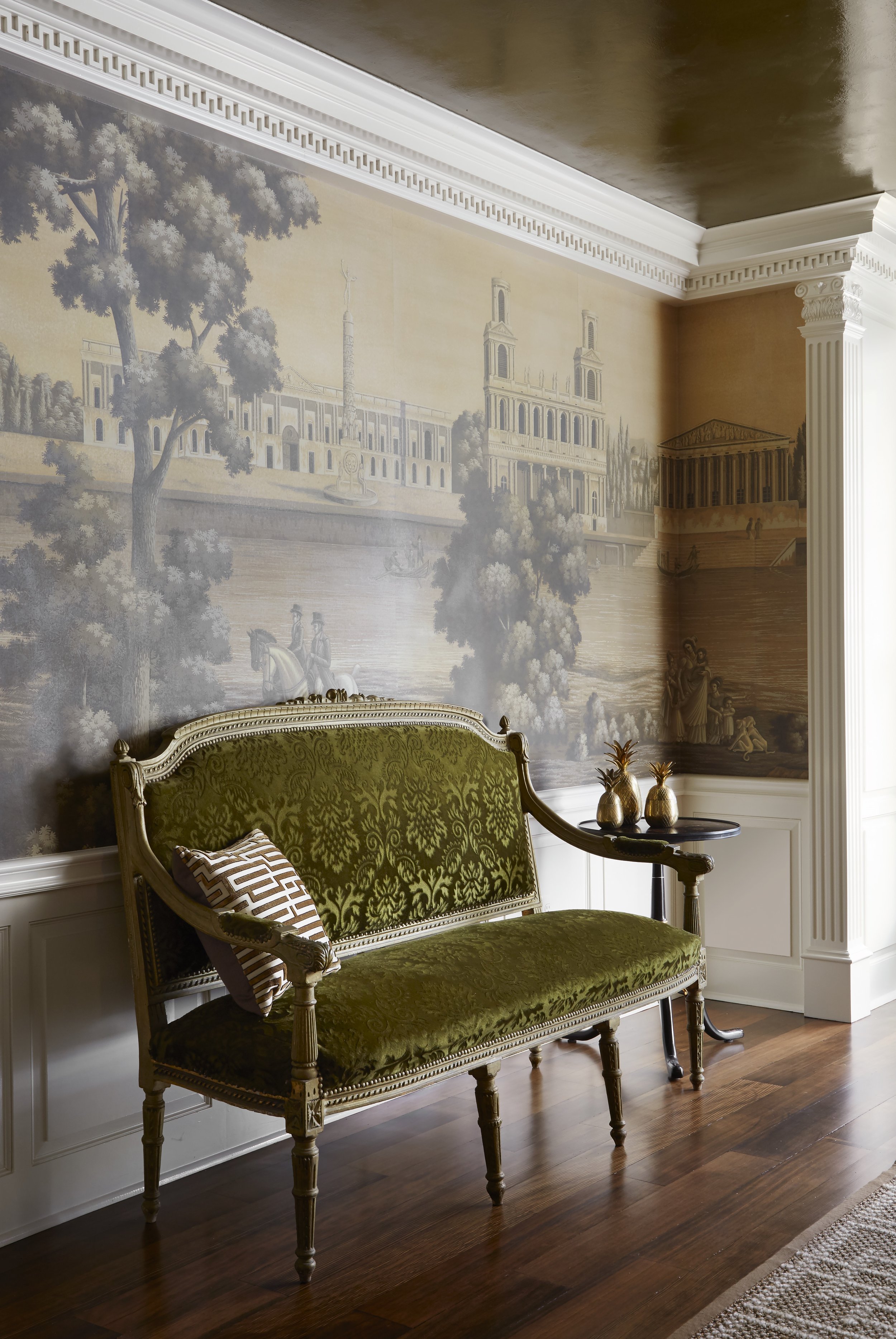 A vintage green velvet upholstered settee with a carved wooden frame and a decorative cushion, set in a room with a mural depicting a historical scene and classical architecture on the wall.