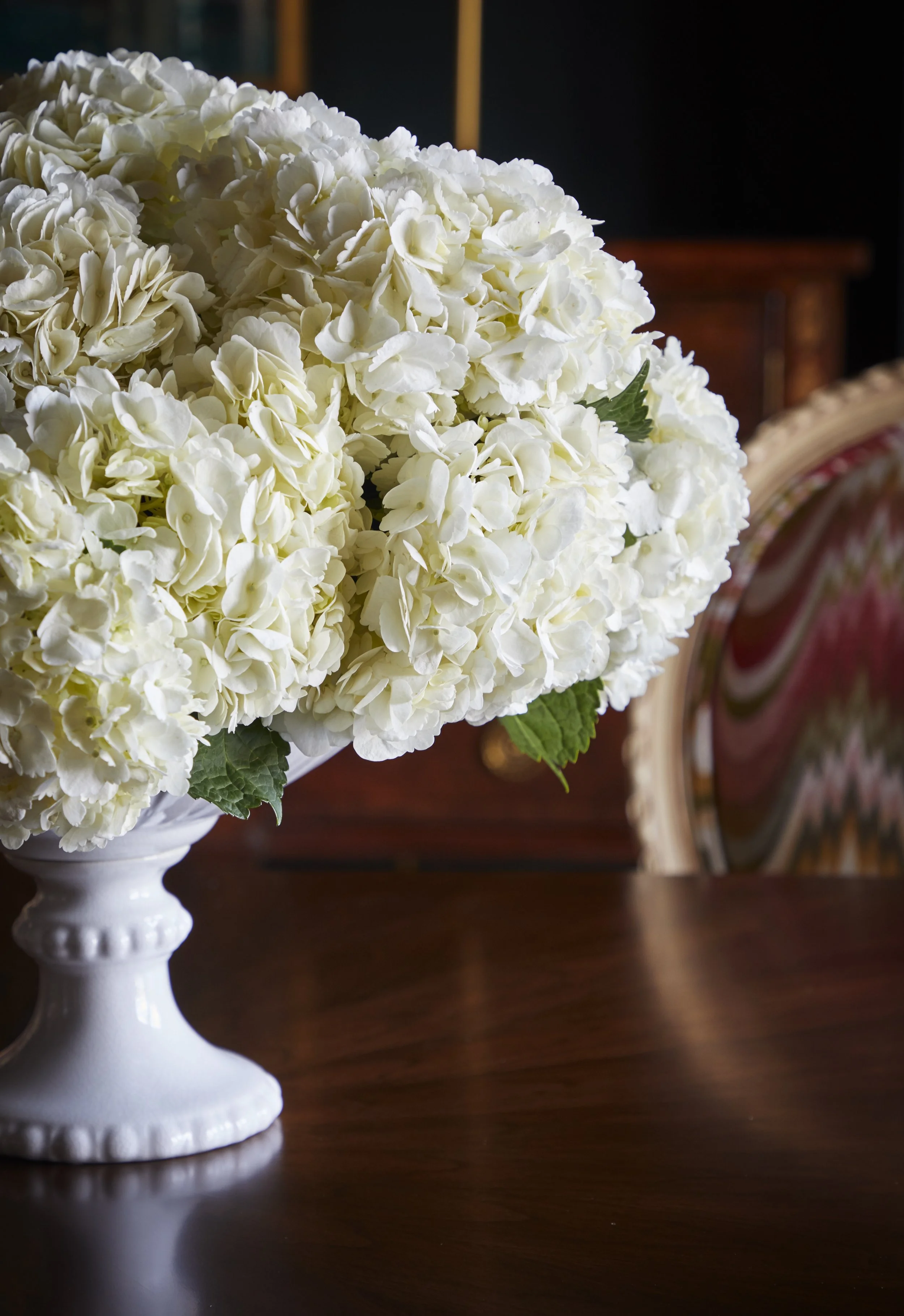 A white ceramic vase holding a large bouquet of white hydrangea flowers on a wooden table, with a blurred background including a wooden cabinet and a partially visible upholstered chair.