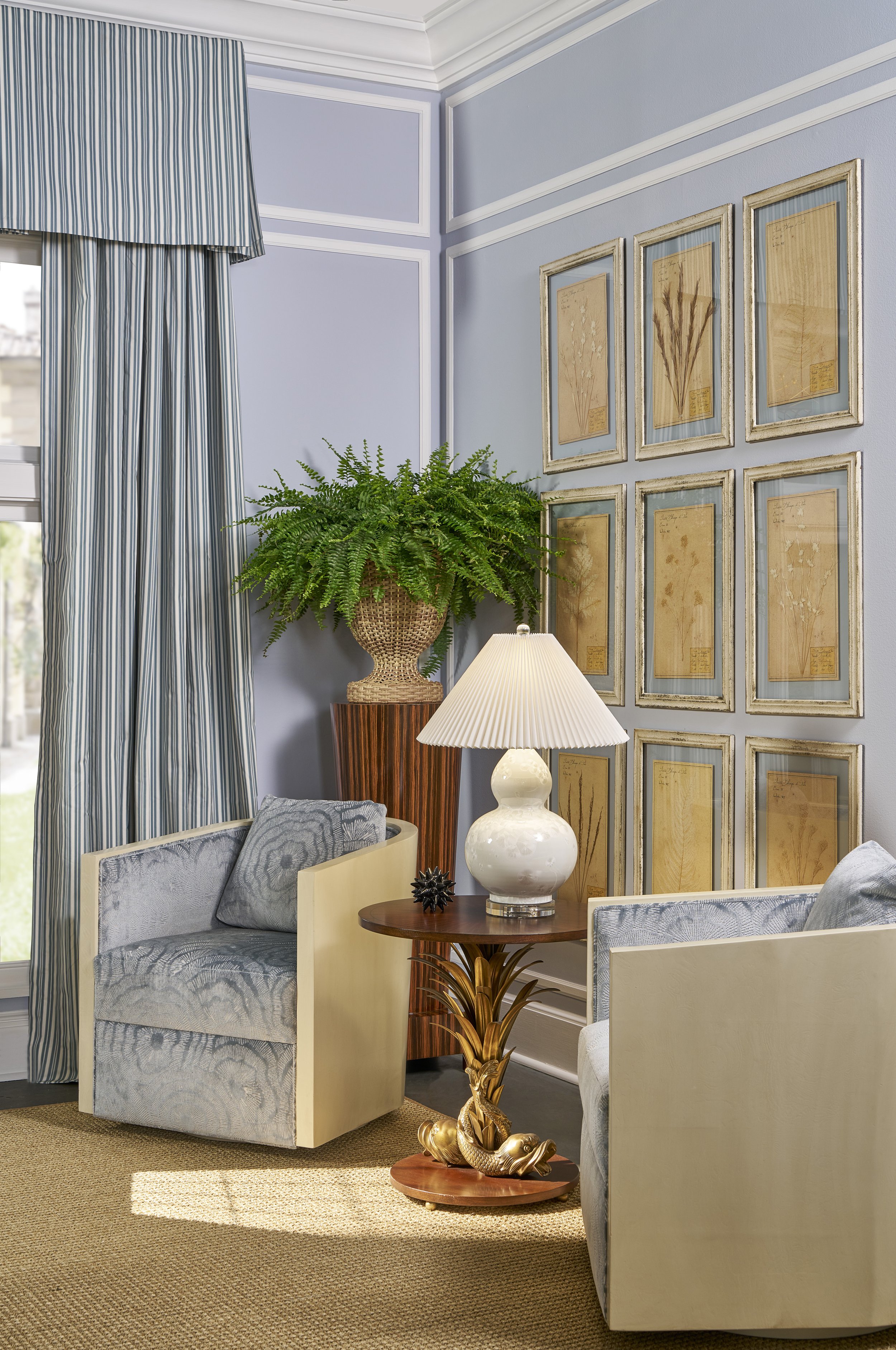 A cozy living room corner with a window adorned with blue and white striped curtains, a potted fern on a wooden stand, a beige armchair with a patterned pillow, a small wooden side table with a white ceramic lamp, and framed botanical prints on the l