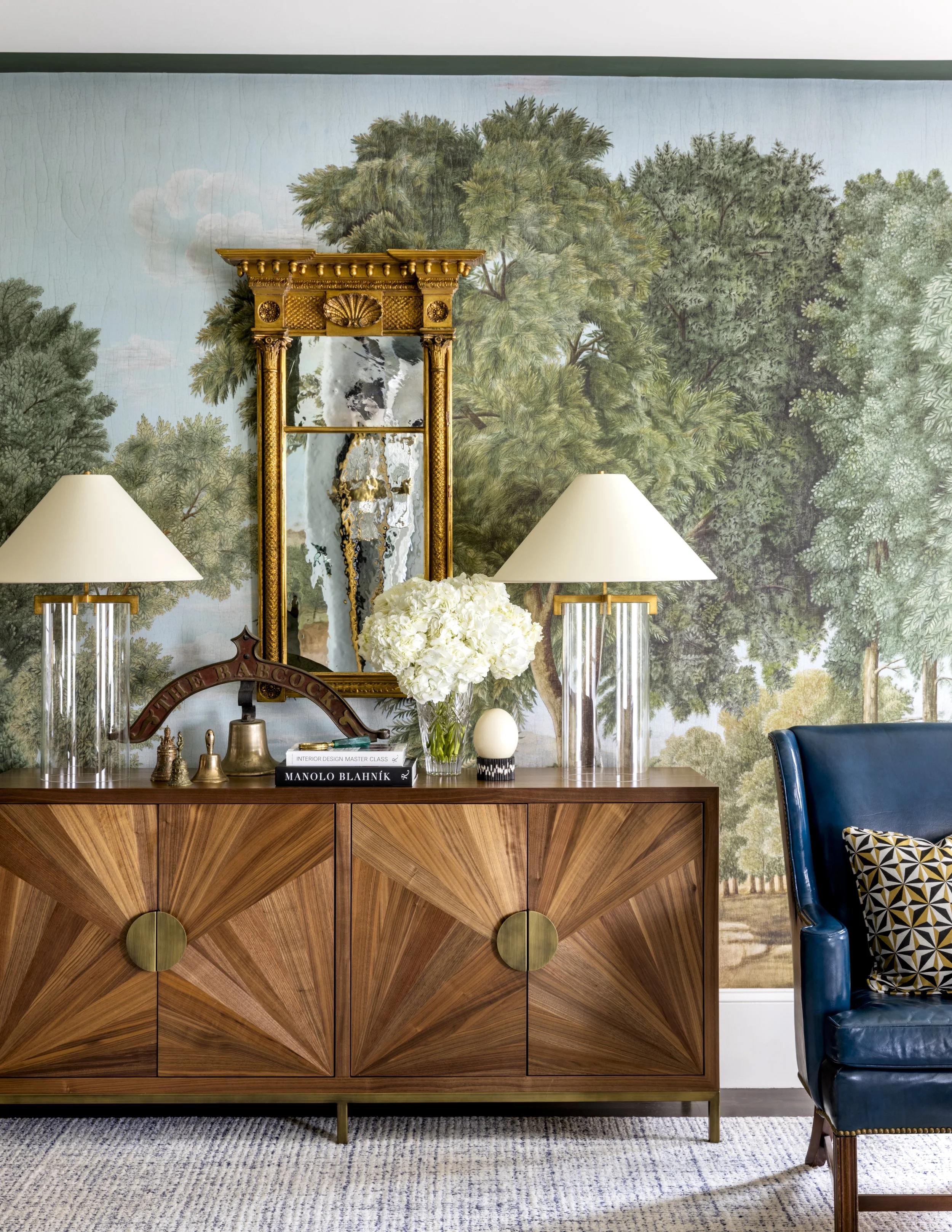 A wooden sideboard with a geometric pattern, a large mirror with ornate gold frame, white flowers in a vase, two tall table lamps with glass bases, and decorative objects on top, in front of a mural of trees and clouds.