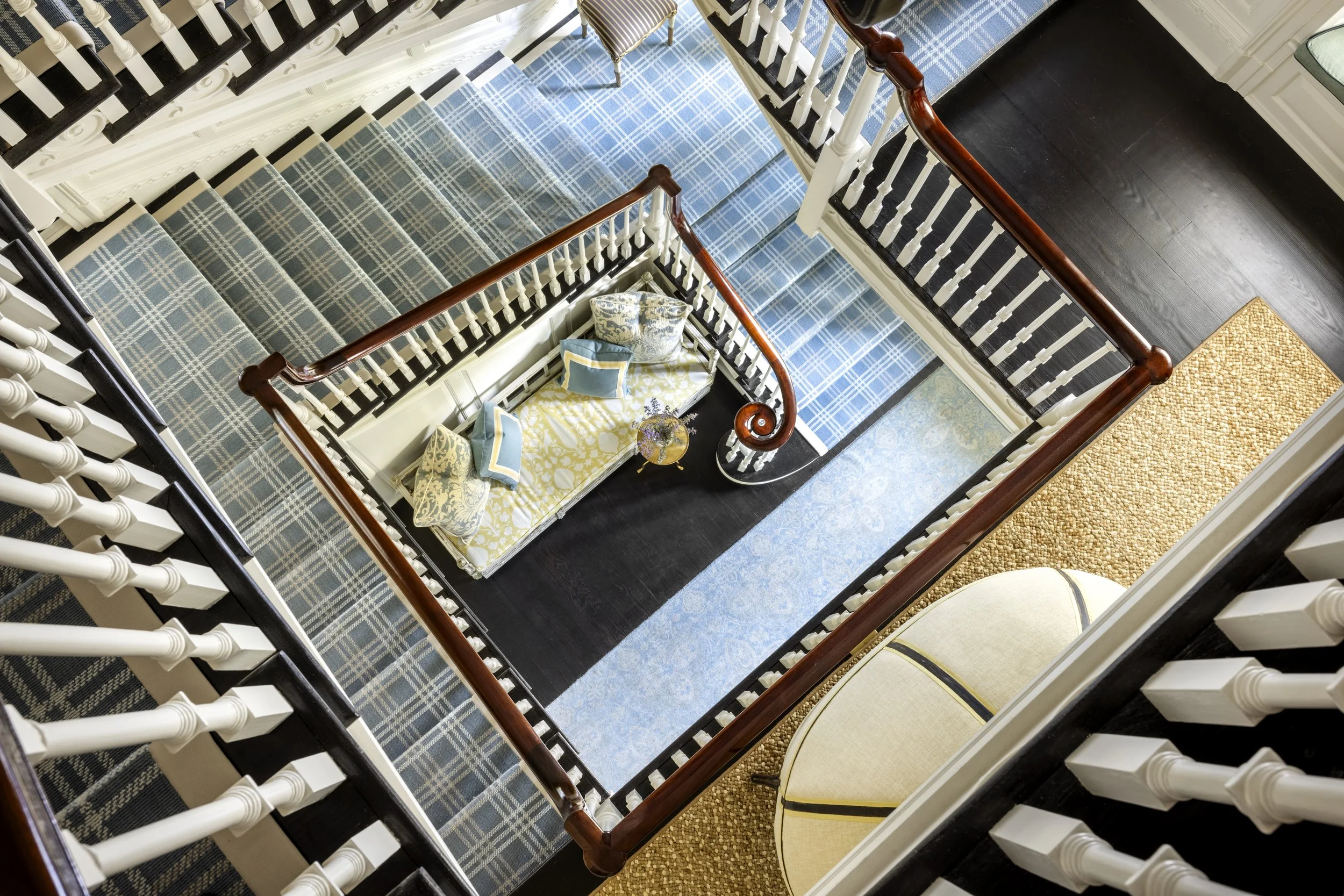 Top view of a staircase with plaid carpet, white spindles, dark and light wood handrails, leading to a sitting area with a yellow patterned sofa, pillows, and a table with flowers.