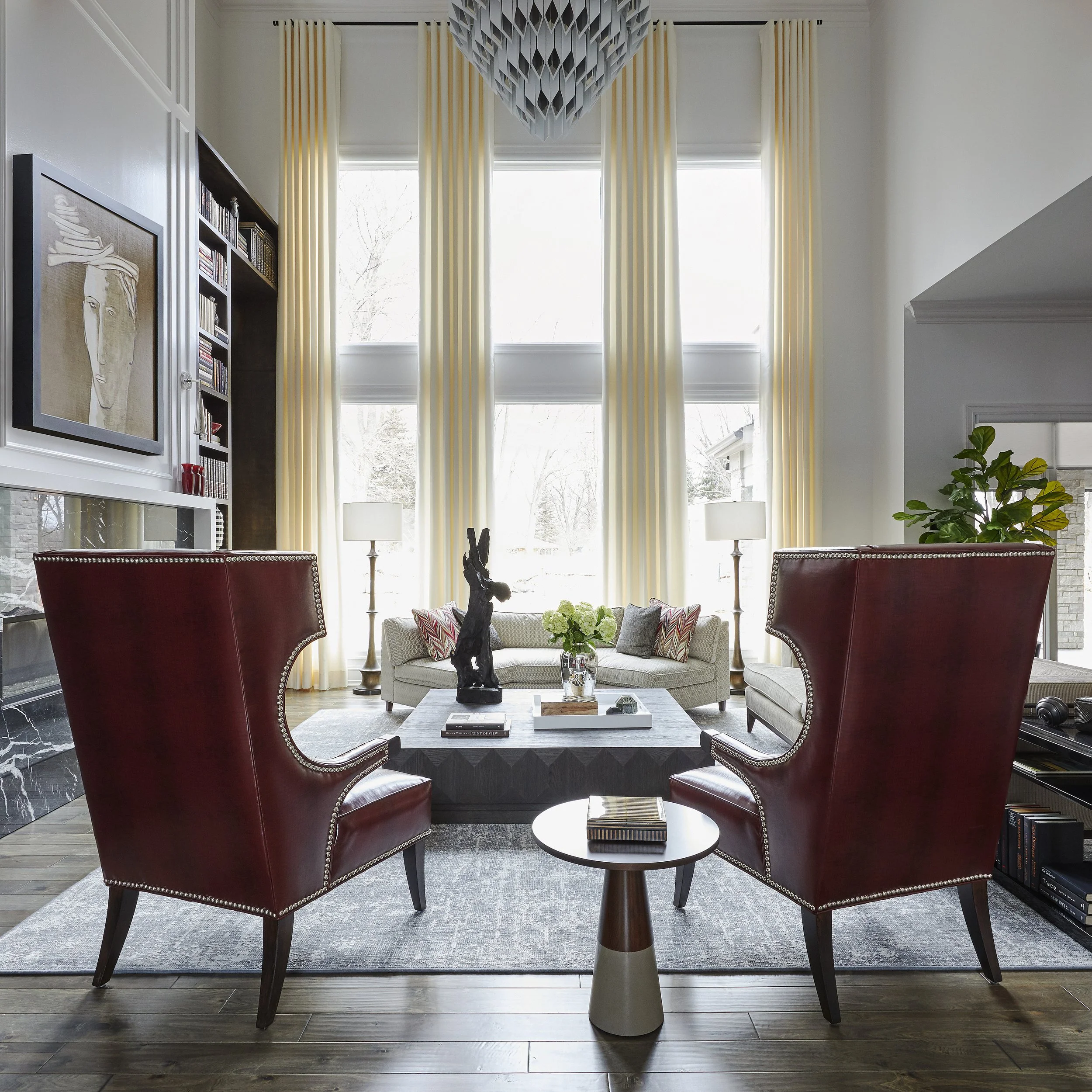 Living room with tall windows, cream curtains, a sculpture on a coffee table, two red chairs, a white sofa with pillows, a bookshelf, art on the wall, and a chandelier hanging from the ceiling.