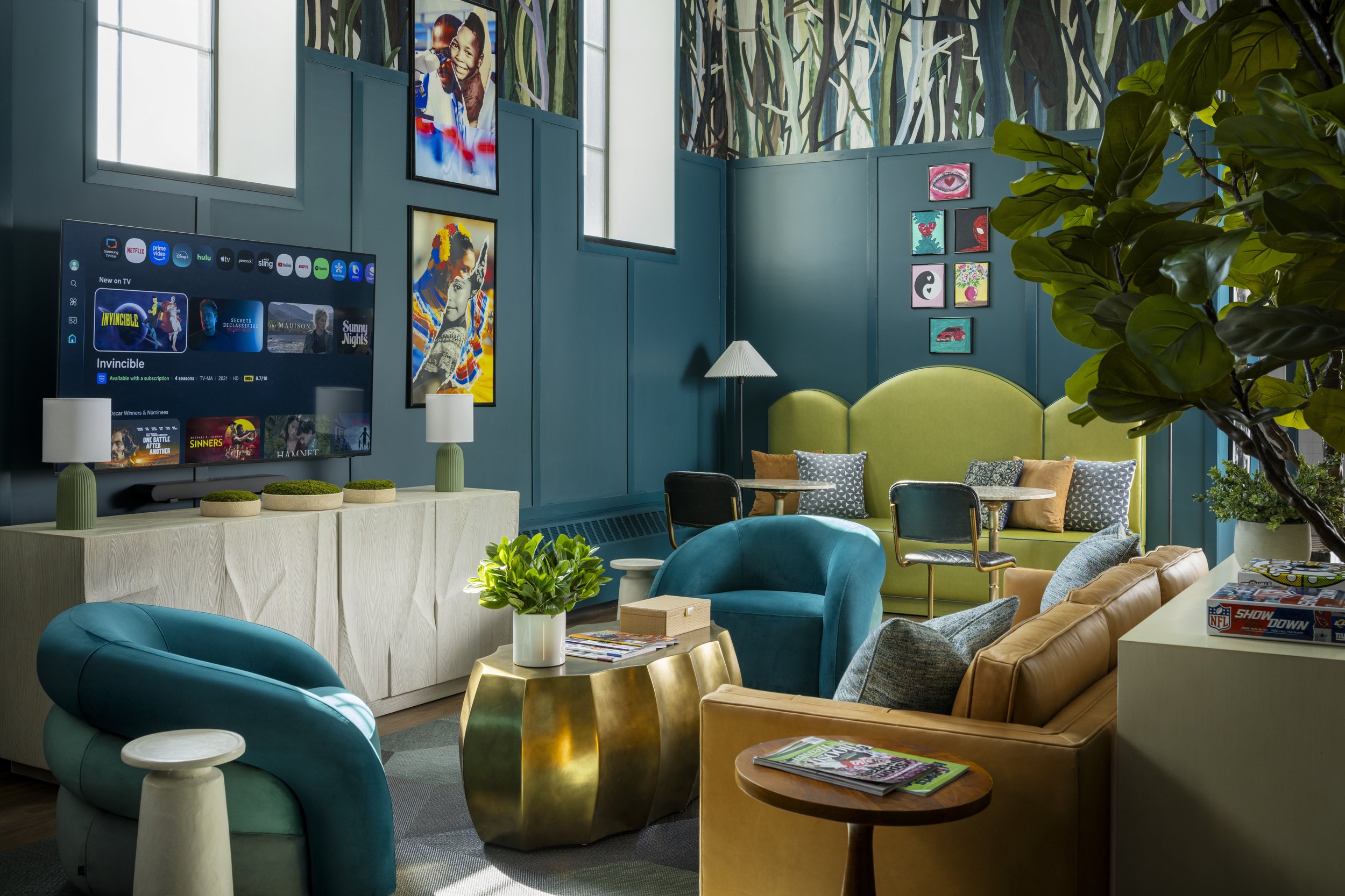 Colorful lounge area with teal, yellow, and blue chairs, framed artwork on teal walls, a potted plant, a flat screen TV displaying streaming services, and a window with a painted mural of a forest scene at the top.