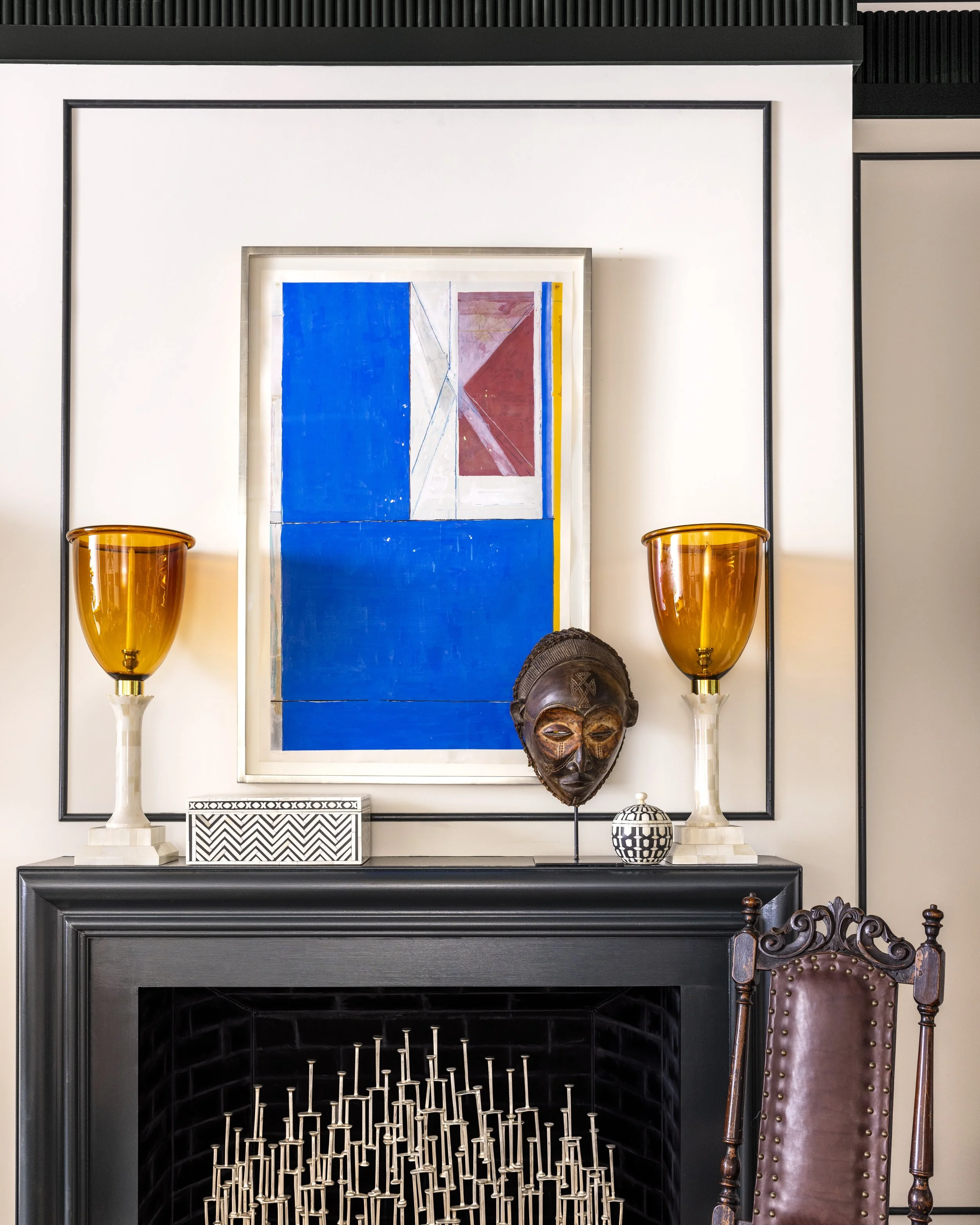 A black fireplace surround with a white mantel, a colorful abstract painting in a silver frame, and two amber glass wall sconces on either side. A carved wooden mask sits on the mantel, and an ornate wooden chair is partially visible in the lower rig