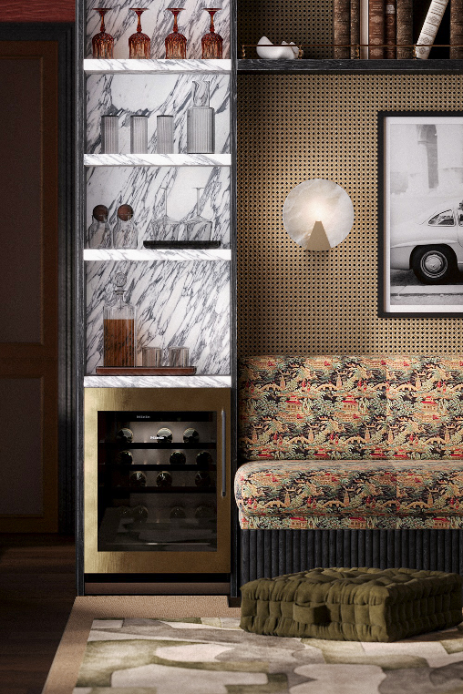 Interior view of a modern living room with a bar area featuring a small wine fridge, decorative shelves with glassware and bottles, a patterned upholstered sofa, a framed black and white photo, a wall-mounted light, and a patterned rug.