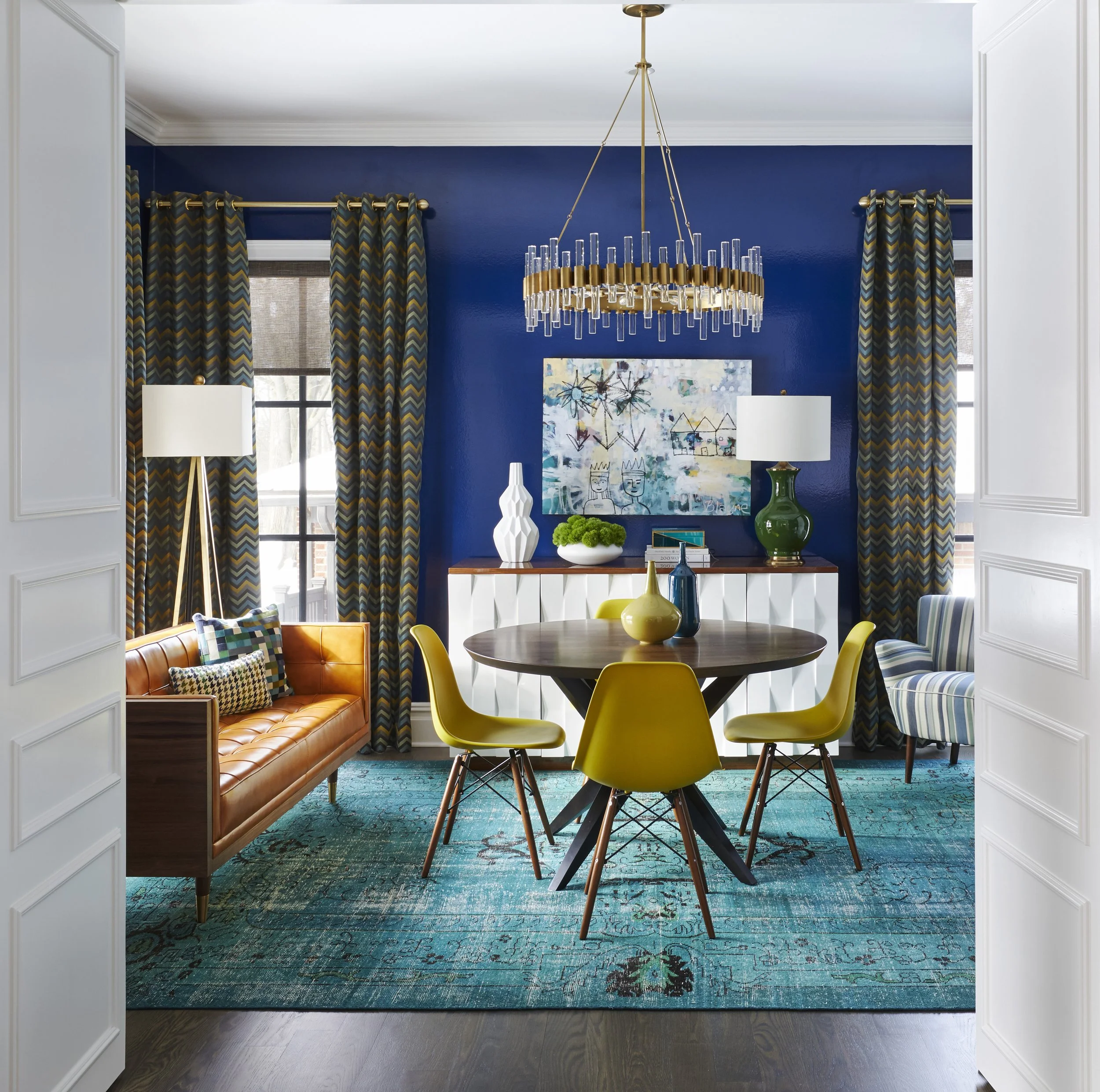 View of a modern dining room with a round wooden table, four yellow chairs, colorful vases, a large abstract painting on a blue wall, patterned curtains, and a glass chandelier hanging from the ceiling.