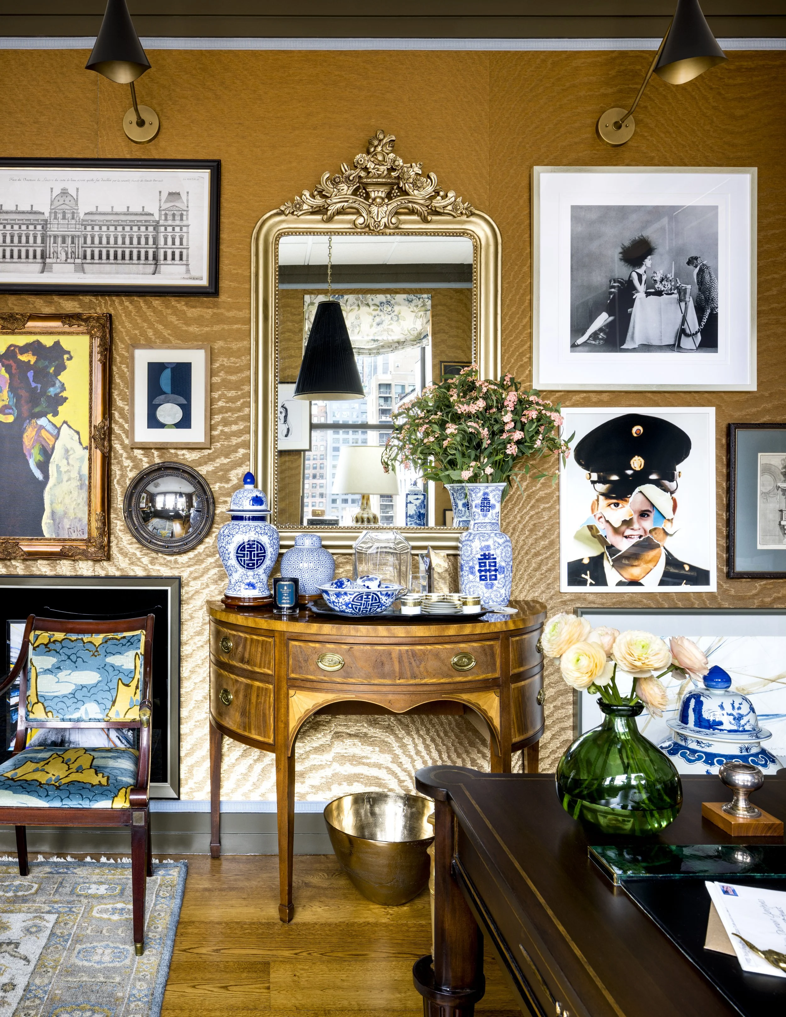 A room decorated with various artworks and a large gold-framed mirror. There is a wooden table with blue-and-white porcelain vases and flowers, as well as framed pictures on the wall, a chandelier reflected in the mirror, and a side table with a gree