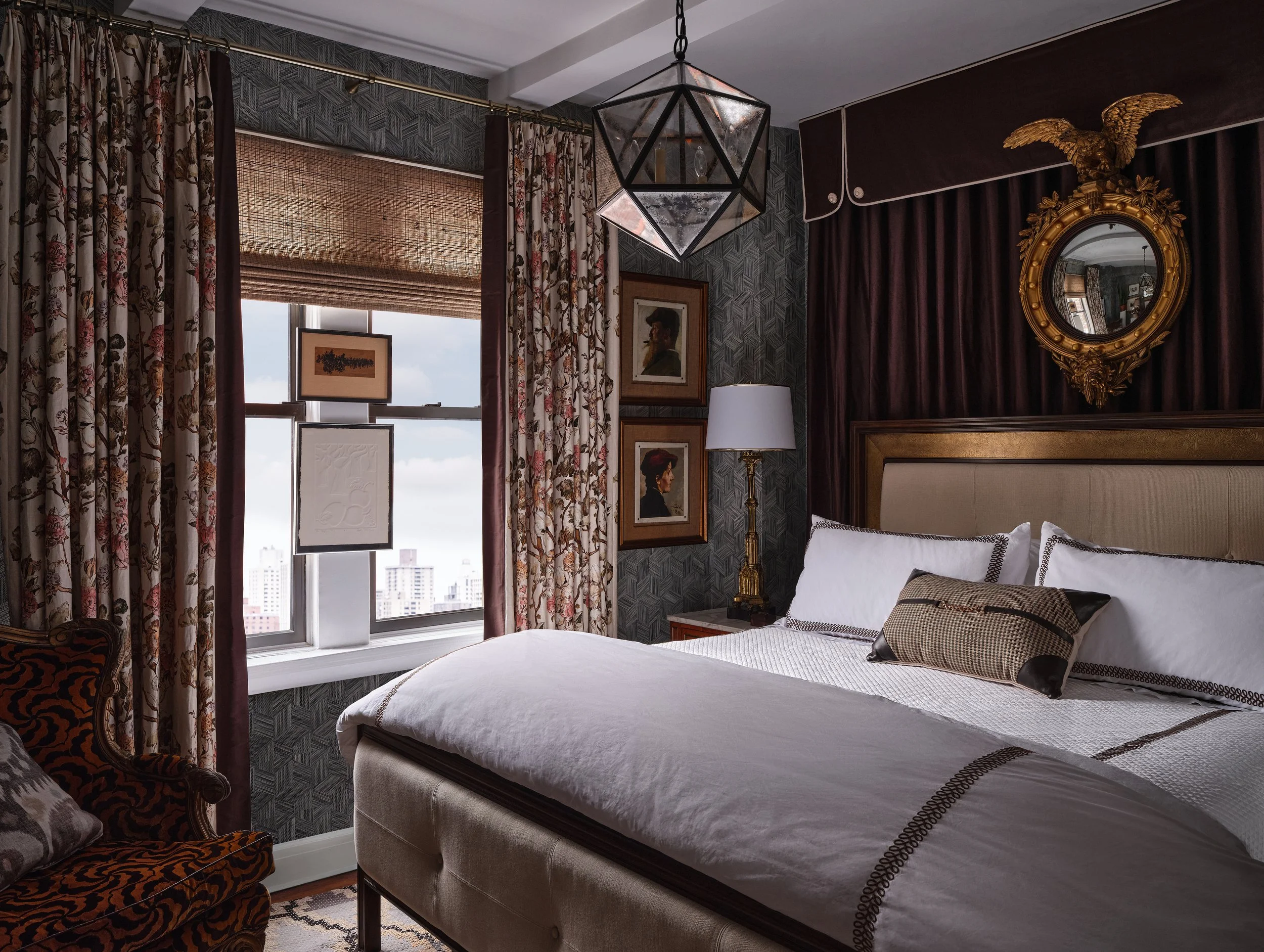 A bedroom with decorated walls, floral curtains, framed artwork, and a bed with white linens and decorative pillows, illuminated by natural light from a window.