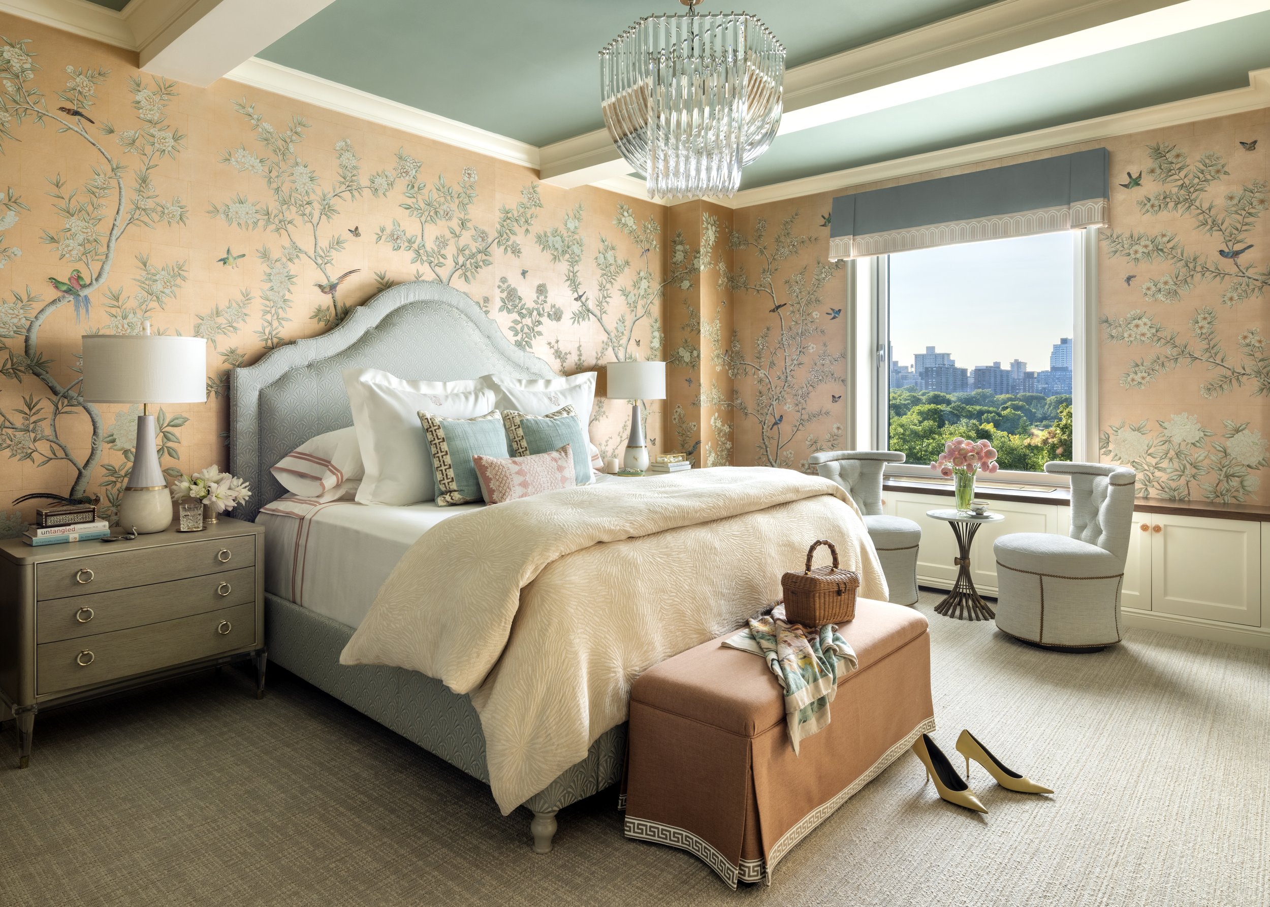 A bedroom with pastel-colored theme featuring a plush bed with various decorative pillows, a tufted beige headboard, and a matching ottoman. The walls have botanical and bird-themed wallpaper, two bedside lamps, a side table with books and flowers, a