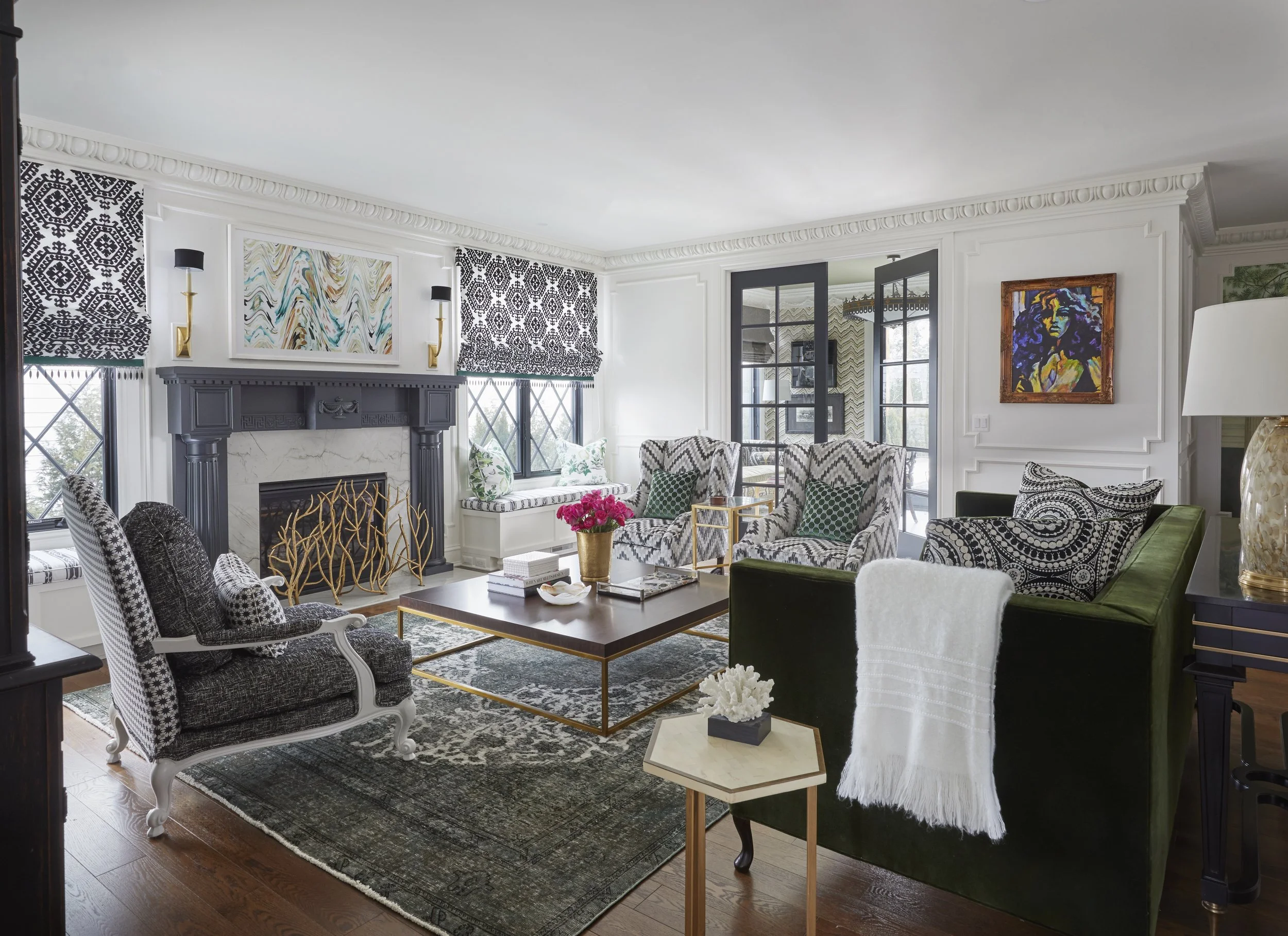 Living room with black, white, and green striped couches, patterned armchairs, artwork above the fireplace, gold side tables, and a vase of pink flowers on a coffee table.