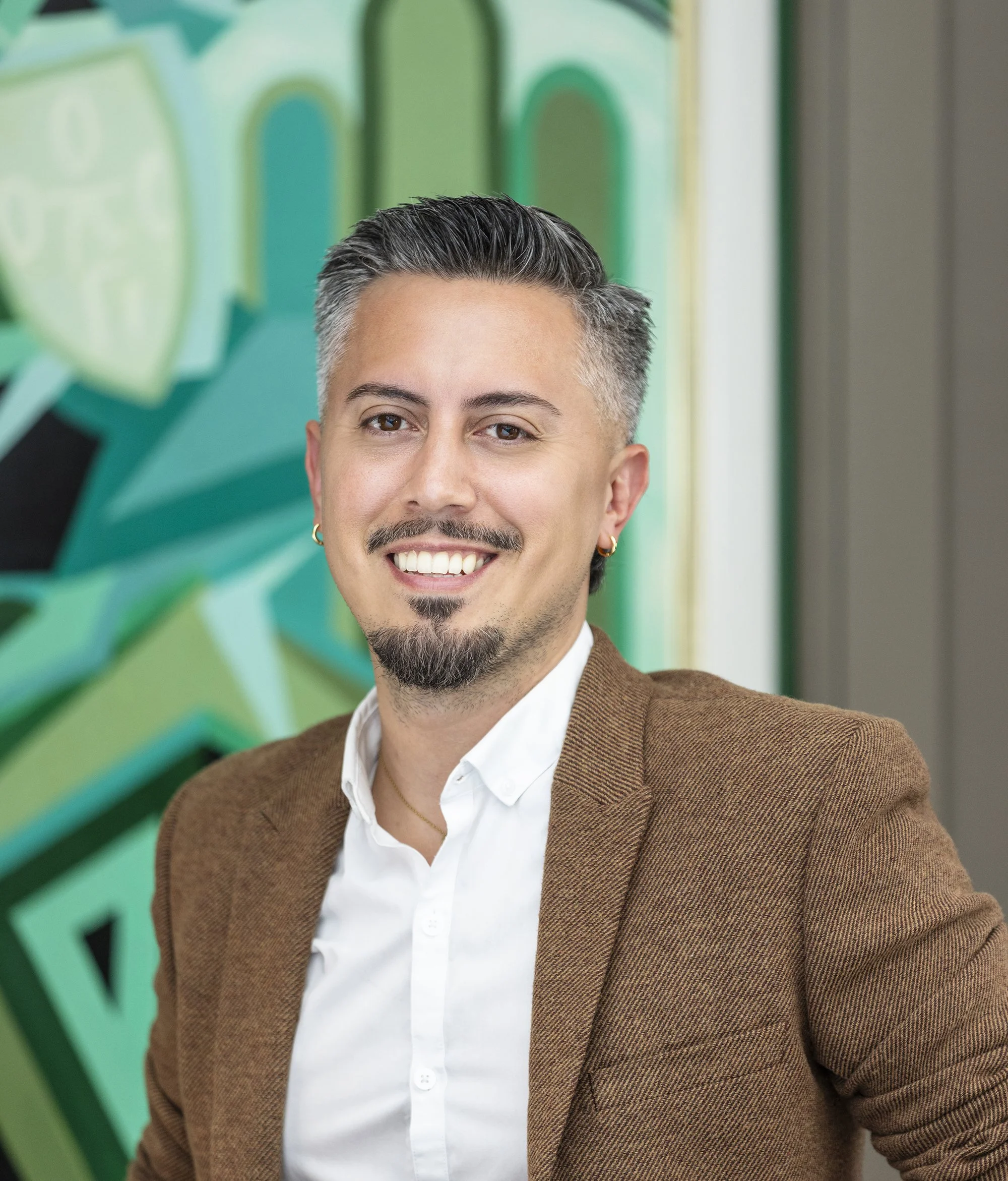 A man with short gray and dark hair, a goatee, and hoop earrings, smiling and wearing a brown blazer and white shirt, standing in front of a colorful mural with green, yellow, and black shapes.