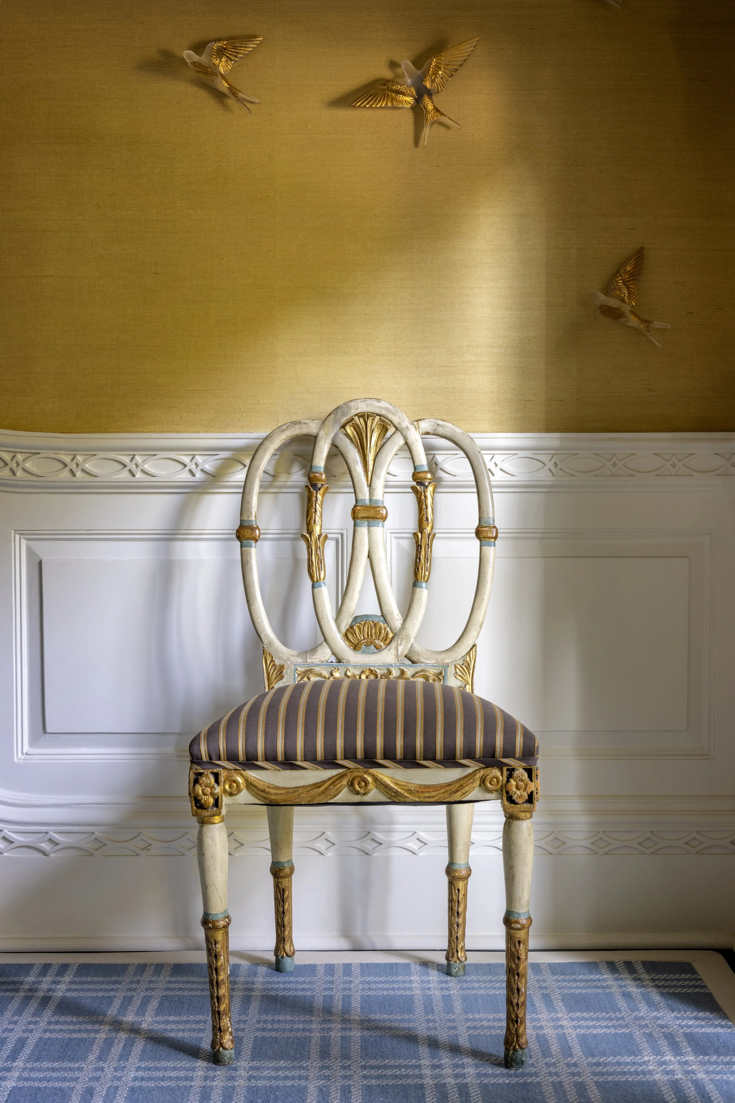 An elaborately decorated cream-colored chair with gold accents and a striped cushion, placed in front of a white paneled wall and a gold wall decorated with three gold and white bird ornaments.