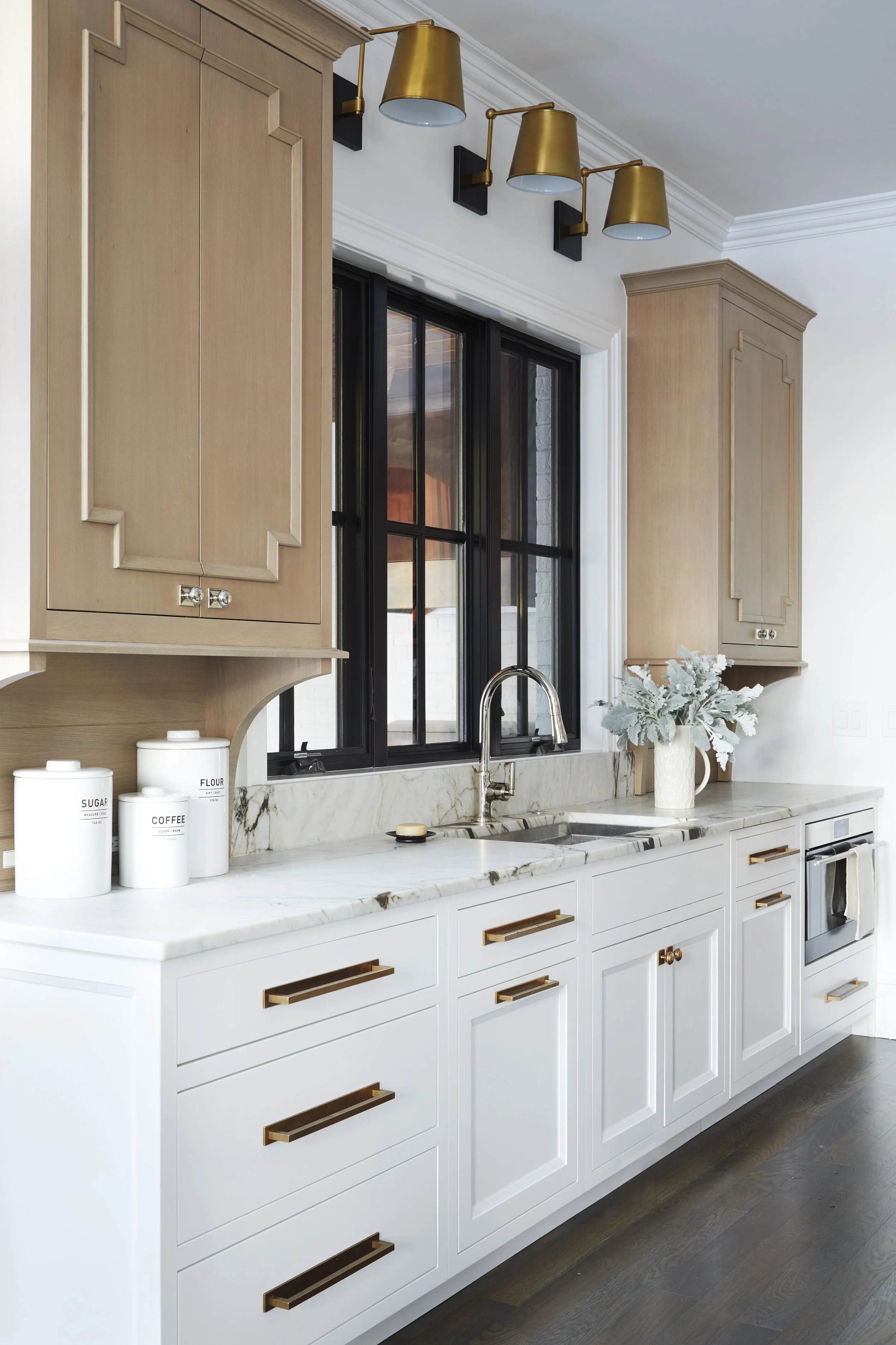 A modern kitchen with white cabinets that have gold handles, a marble countertop, a stainless steel sink, and black-framed windows. Above the window, there are three gold wall sconces with black bases, and to the side, there are light wooden cabinets