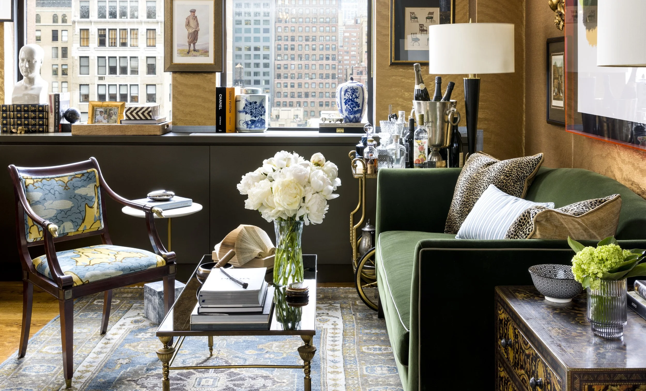 Interior view of a luxurious living room featuring a green sofa with decorative pillows, a glass coffee table with a large flower arrangement, patterned armchair, books, artwork on the walls, and tall windows with a cityscape view.