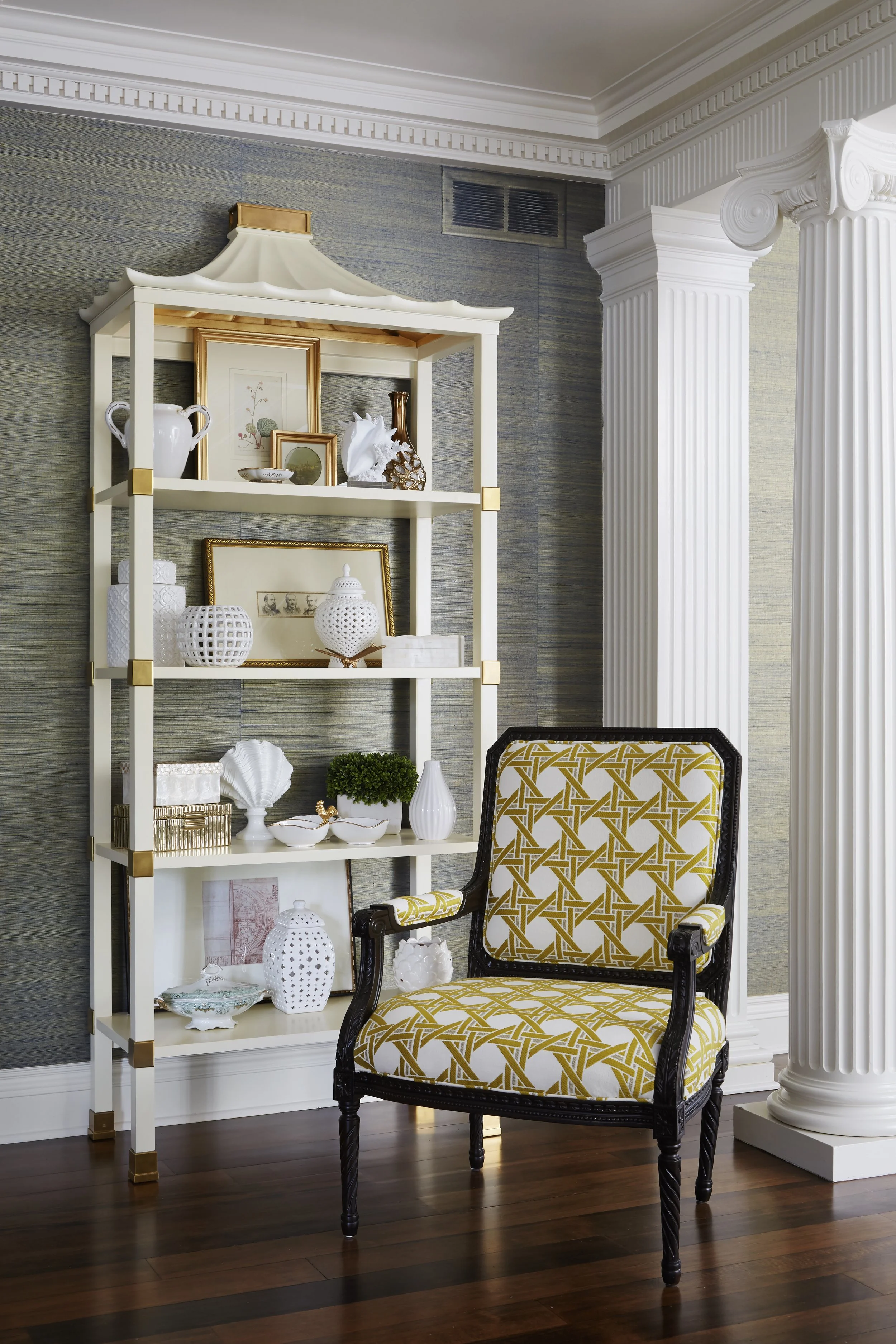 Decorative interior with a white shelf holding ceramic and framed artwork, a yellow and white patterned armchair, and classical columns.
