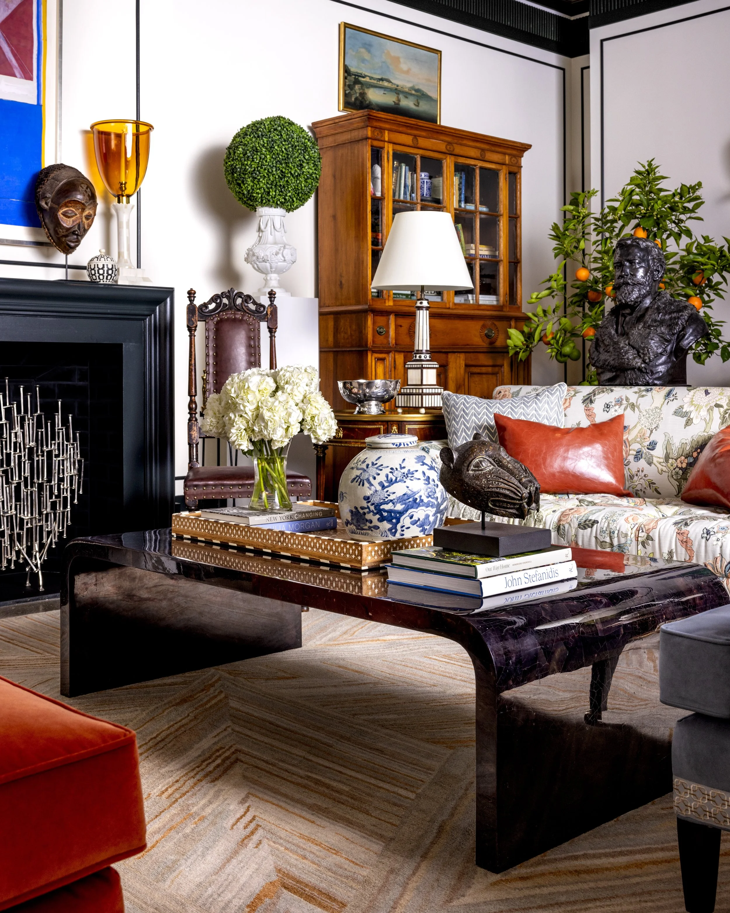 Living room with a black marble coffee table, floral-patterned sofa with cushions, a bouquet of white hydrangeas, a decorative brass or bronze bird sculpture, books, a white table lamp, a wooden display cabinet, and a bust sculpture of a man with a b