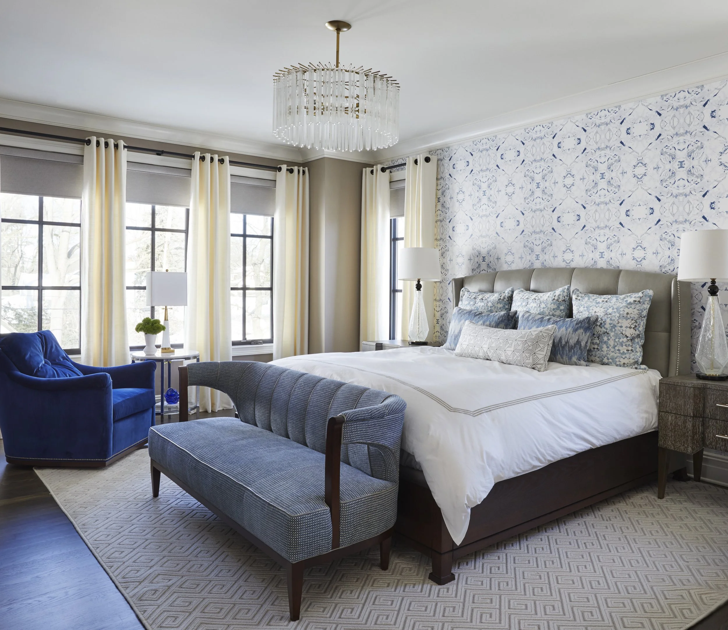 Bedroom with large painted wall, white curtains, window with black grid, bed with white bedding and pillows, blue armchair, side table with lamps, wooden benches at foot of bed, geometric patterned rug, and modern chandelier.