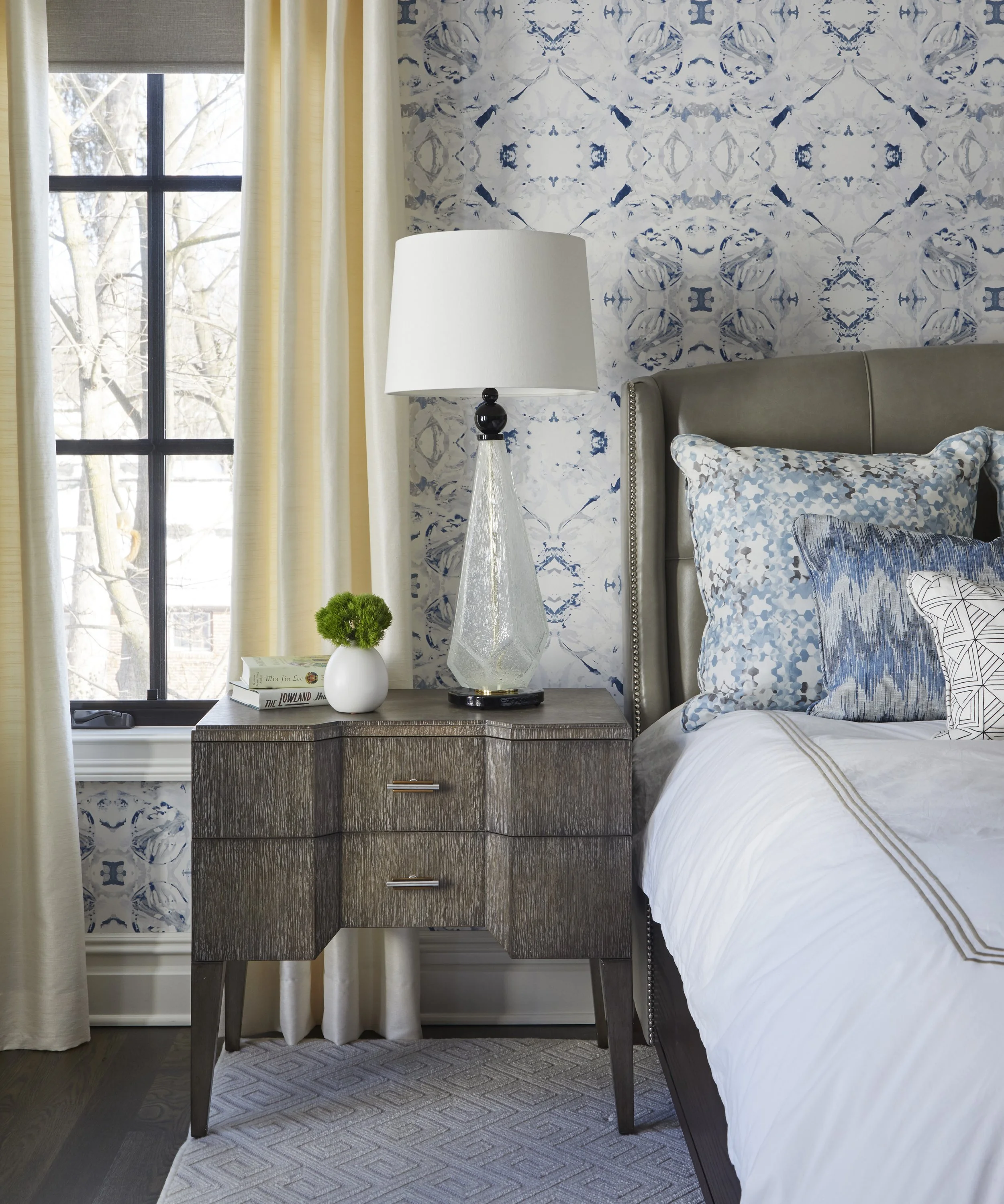 A bedroom corner featuring a bed with patterned pillows, a nightstand with a small green plant, several books, a large decorative glass lamp with a white lampshade, a window with beige curtains, and a wall with blue and white patterned wallpaper.