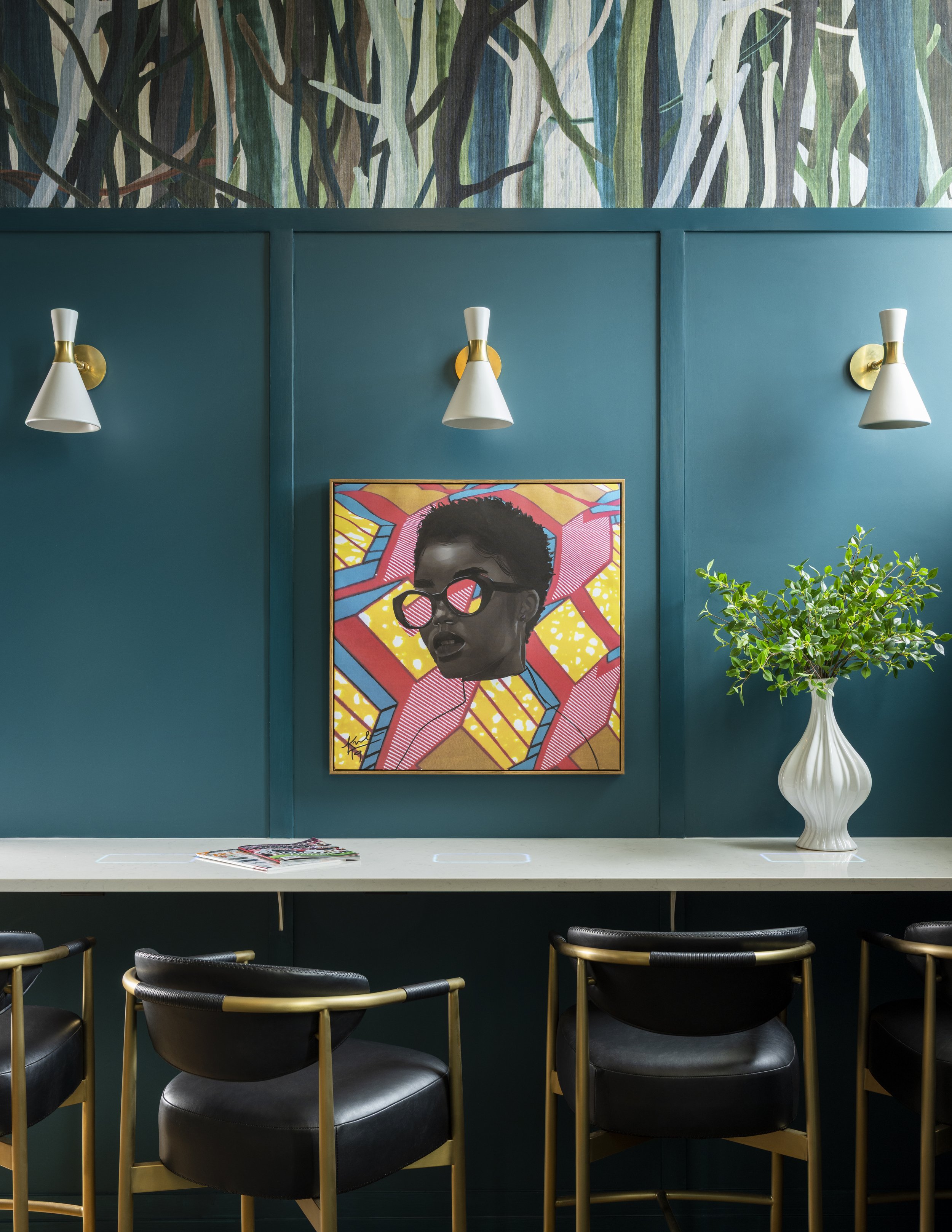 A modern interior space with a teal wall, three white and gold wall sconces, a colorful artwork of a woman with glasses, a white countertop with magazines, a white vase with a green plant, and black chairs with gold accents.