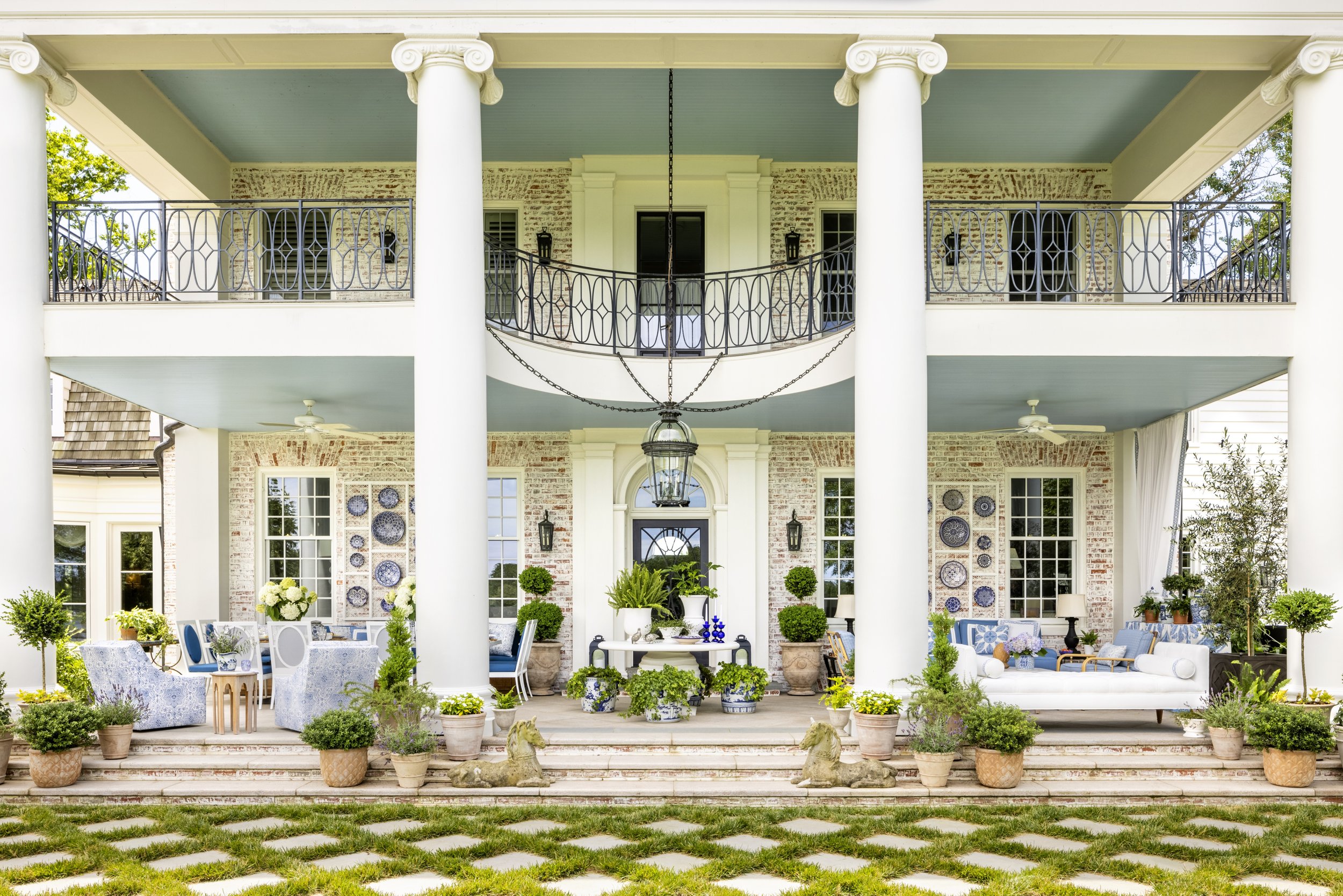 The image shows the front view of a grand house with a porch supported by large white columns. The house features brick walls, large windows, and decorative wall art. The porch is decorated with potted plants, flowers, and outdoor furniture, creating