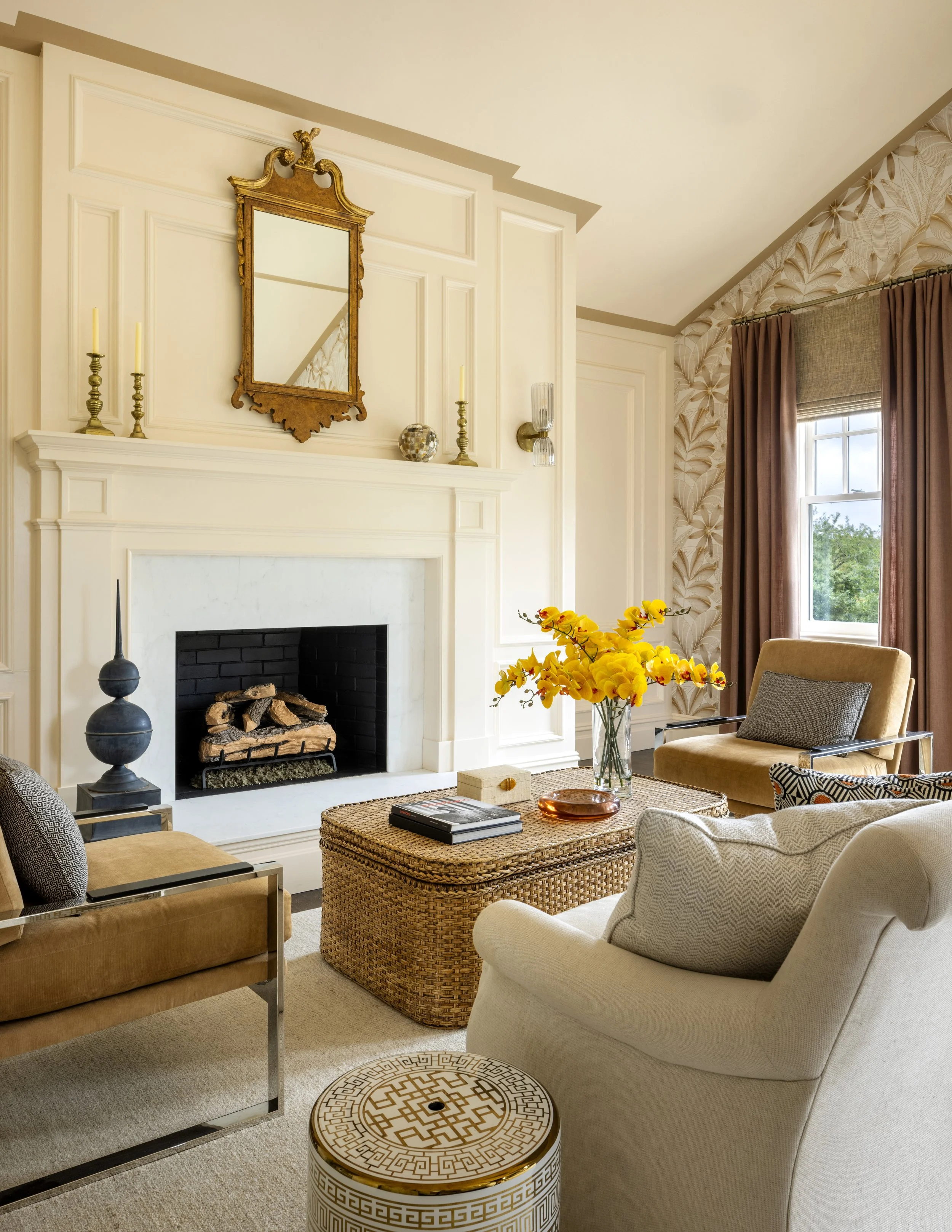 A cozy living room with a fireplace, a mirror above the mantel, and various decorative items including candlesticks, a patterned vase, and a glass container. A large window with drapes and a patterned wallpaper panel are visible. There are sofas and 