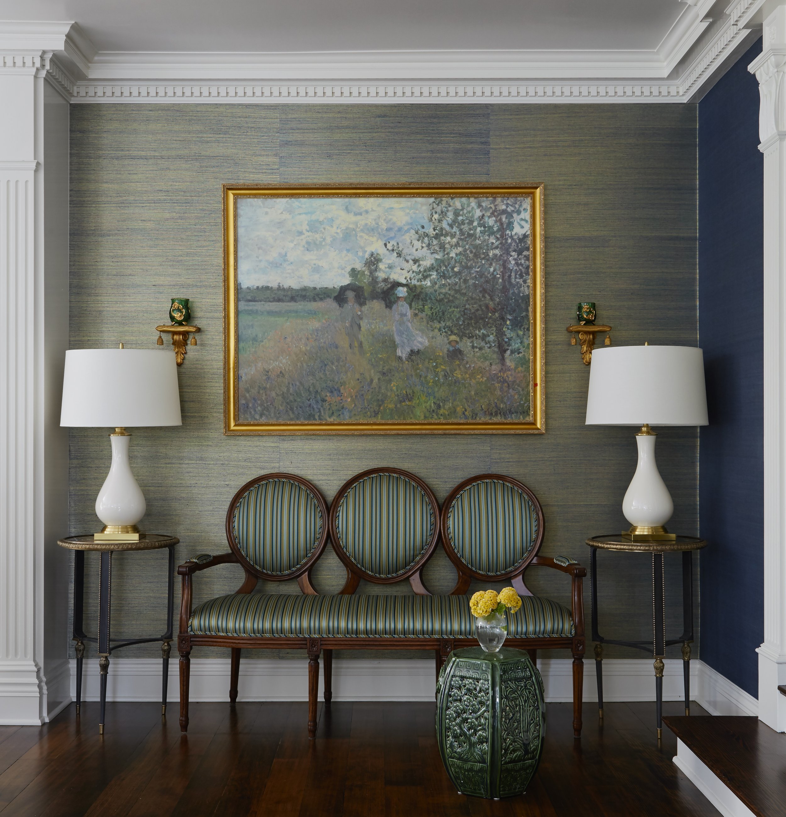 An interior wall with framed artwork, two ivory lamps with gold accents on side tables, a green decorative vase with yellow flowers on a small green table, and chairs with striped fabric seats.