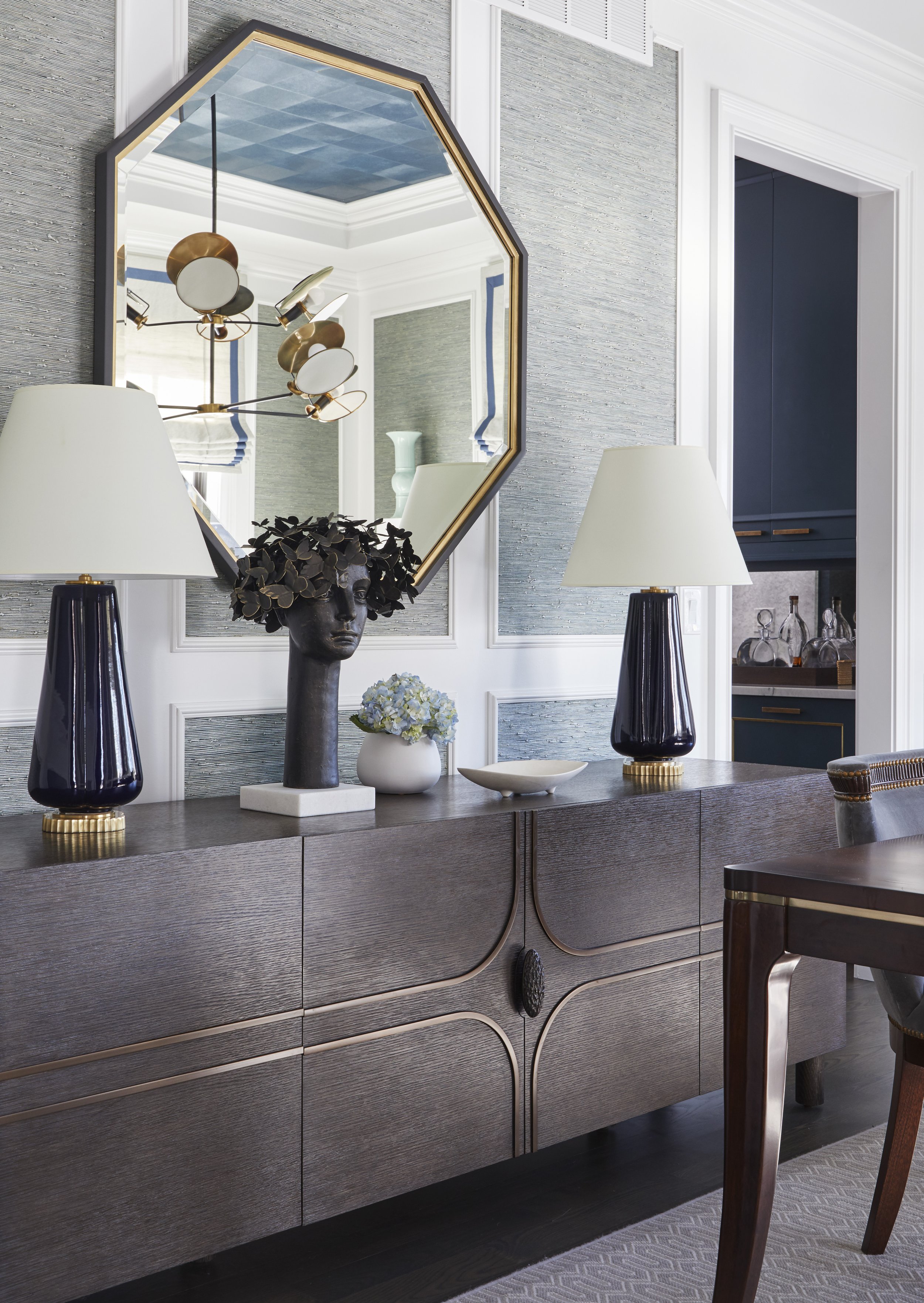 A modern wooden sideboard with a dark finish, decorated with black and gold vases, a statue head with black leaves on top, a white flowerpot with flowers, and a large geometric mirror reflecting a chandelier and part of the room.