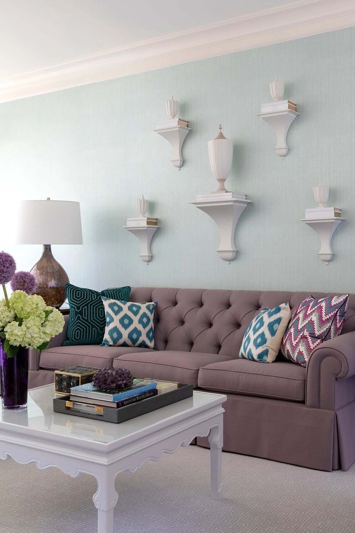 A living room with a pink tufted sofa decorated with blue, white, and pink patterned pillows. A white coffee table has a tray with books and purple flowers. Purple flowers in a vase and a lamp with a brown base are on a side table. Green and purple h