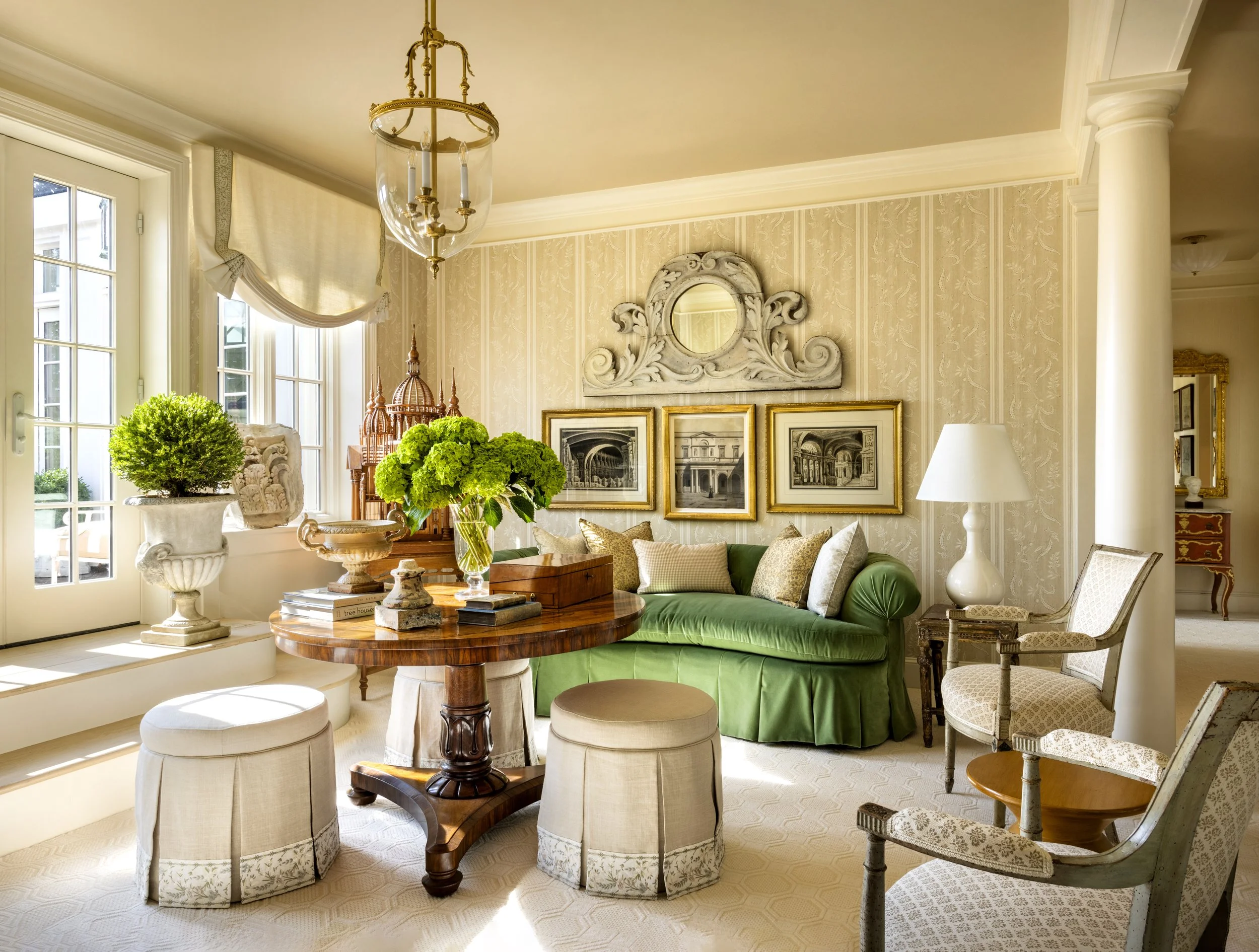 A cozy living room with large windows allowing natural light, a green velvet couch with various pillows, a round wooden table with decorative items and a vase of green hydrangeas, framed artwork on the wall, a white ornate mirror, and a mix of classi