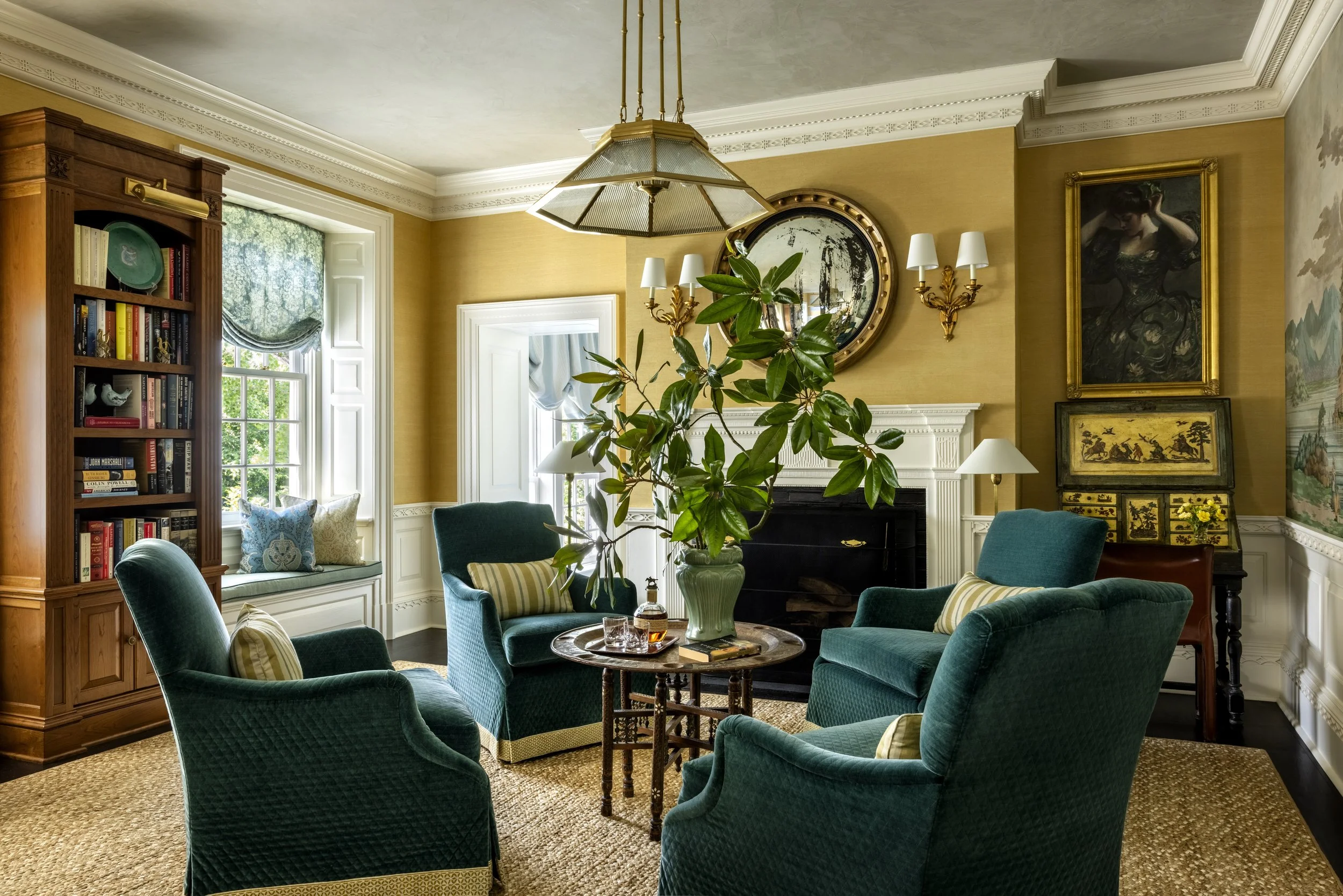 A cozy living room with green upholstered chairs arranged around a wooden coffee table, a large leafy potted plant, built-in bookshelves, and framed artwork on yellow walls, with a large window letting in natural light.