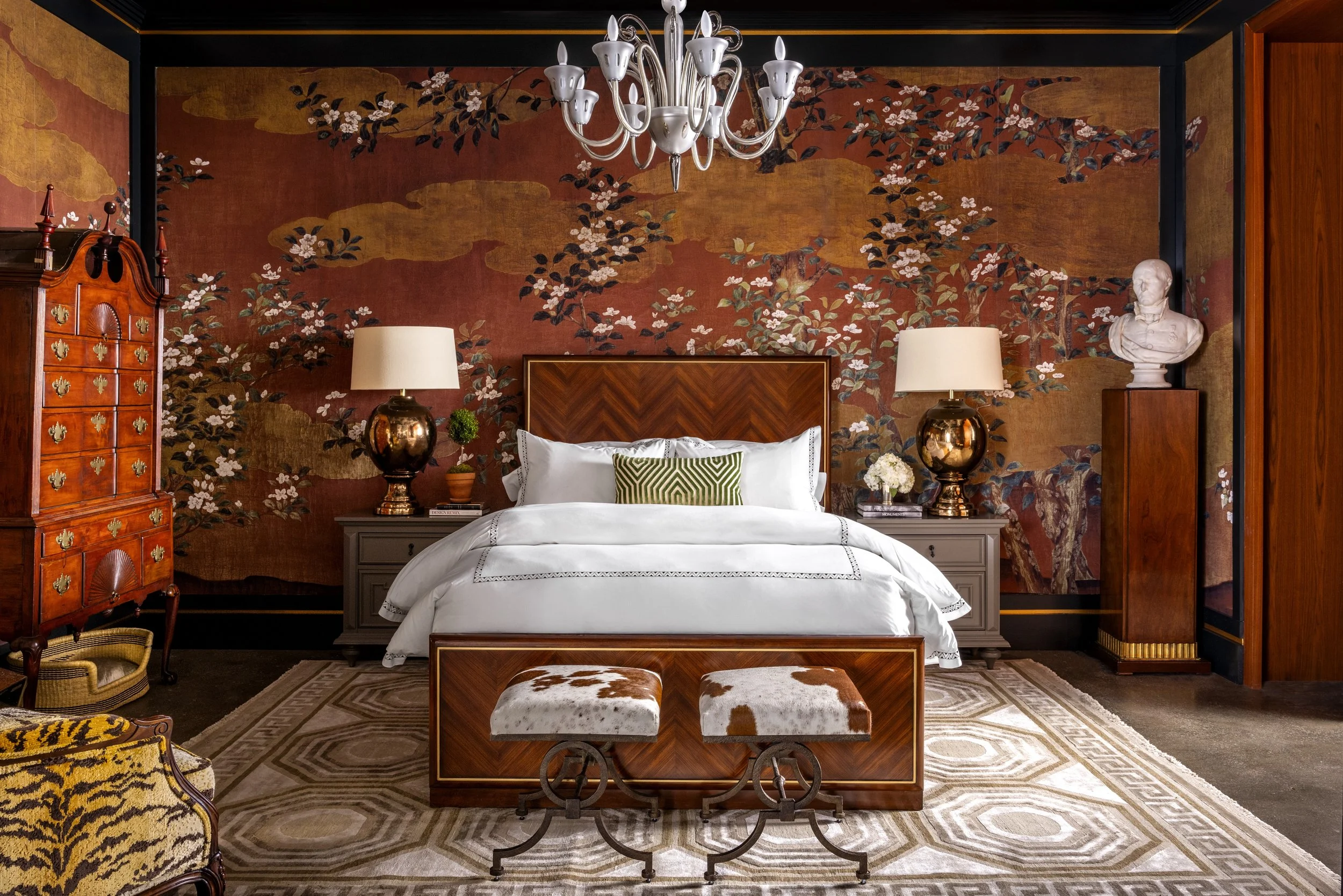 A bedroom with a large bed, a wood headboard, and a decorative floral mural on the wall behind it. There are two nightstands with matching lamps, and a chandelier hanging from the ceiling. The room also features two upholstered stools with cowhide se